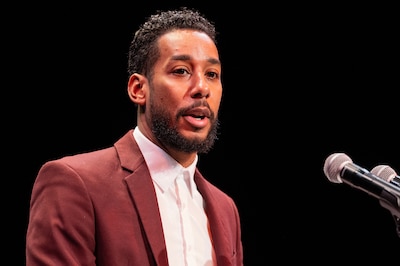 An adult in a red suit jacket speaks from behind a microphone and in front of a red background.