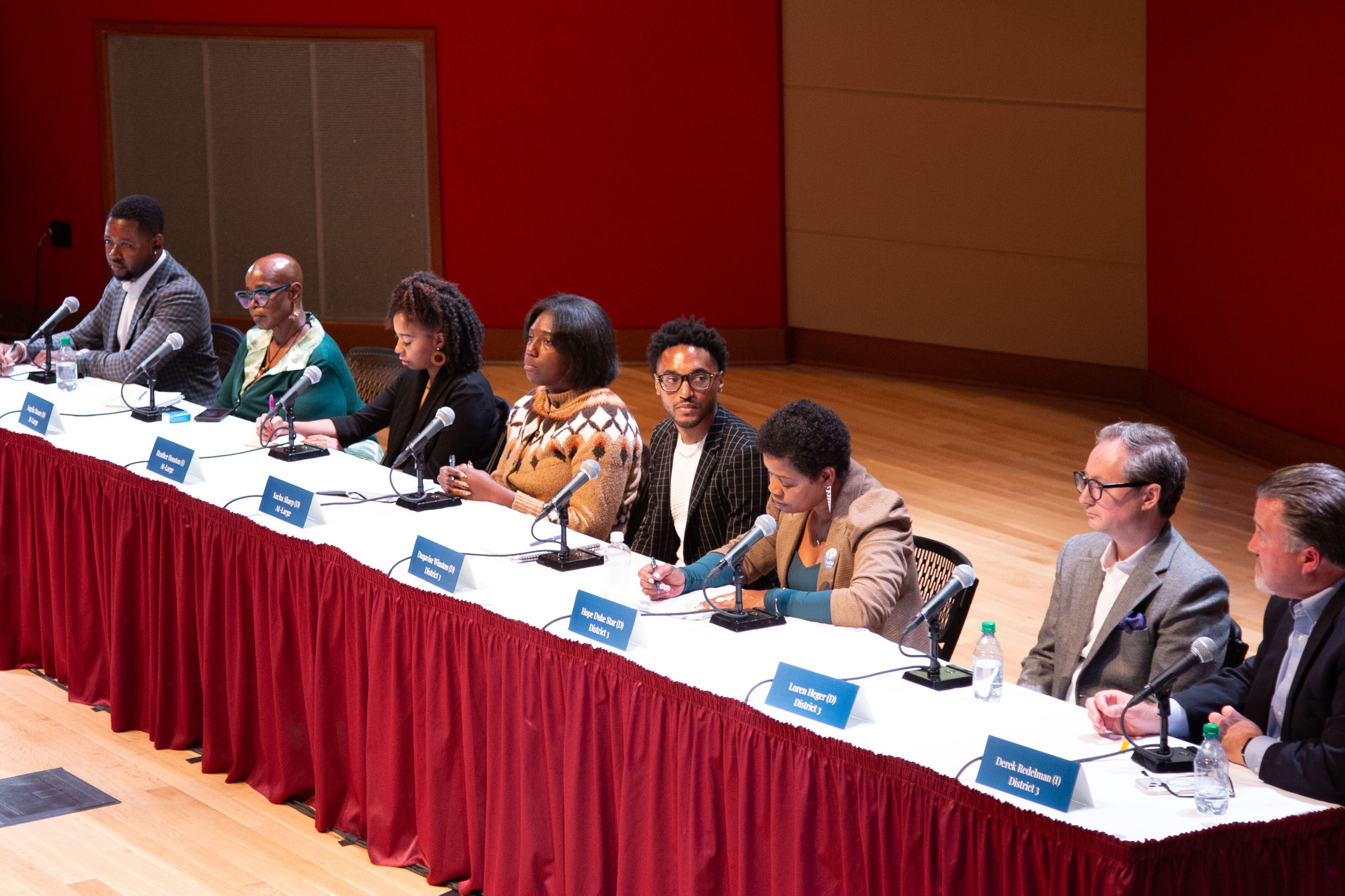 Eight adults sit side by side at a long table during a candidate forum in a large meeting hall.