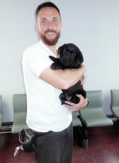 A man wearing a white shirt holds a black dog.