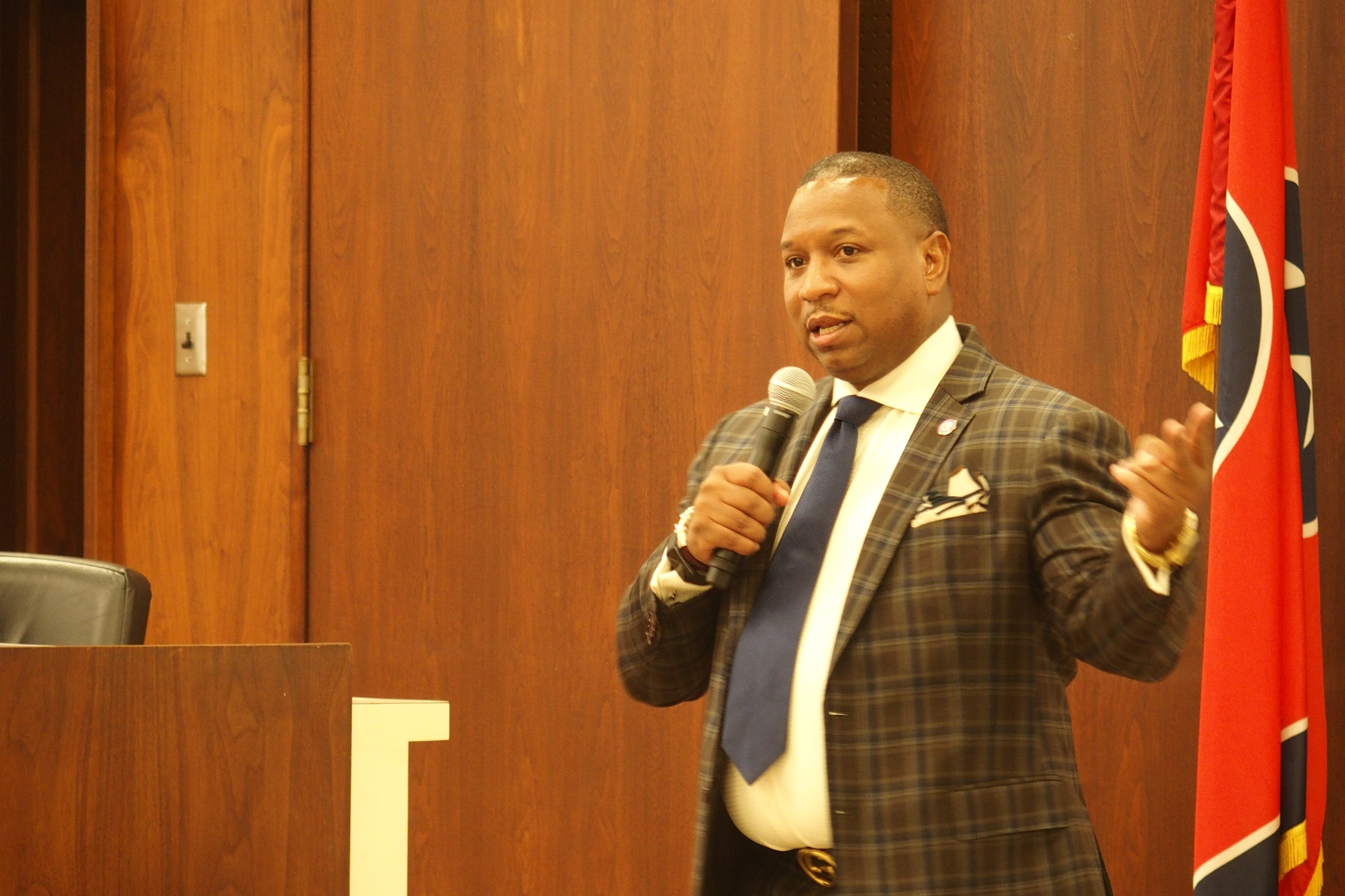 A man wearing a suit speaks into a microphone and as he gestures with his left hand.