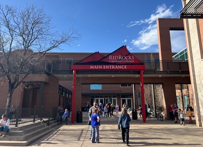 People walk on a concrete sidewalk near large buildings and a red sign that says "Red Rocks Community College" with a blue sky in the background.