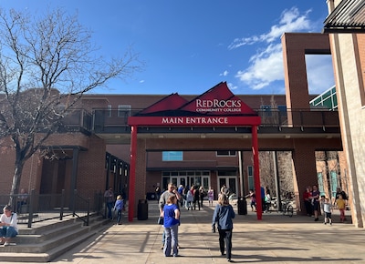 People walk on a concrete sidewalk near large buildings and a red sign that says "Red Rocks Community College" with a blue sky in the background.