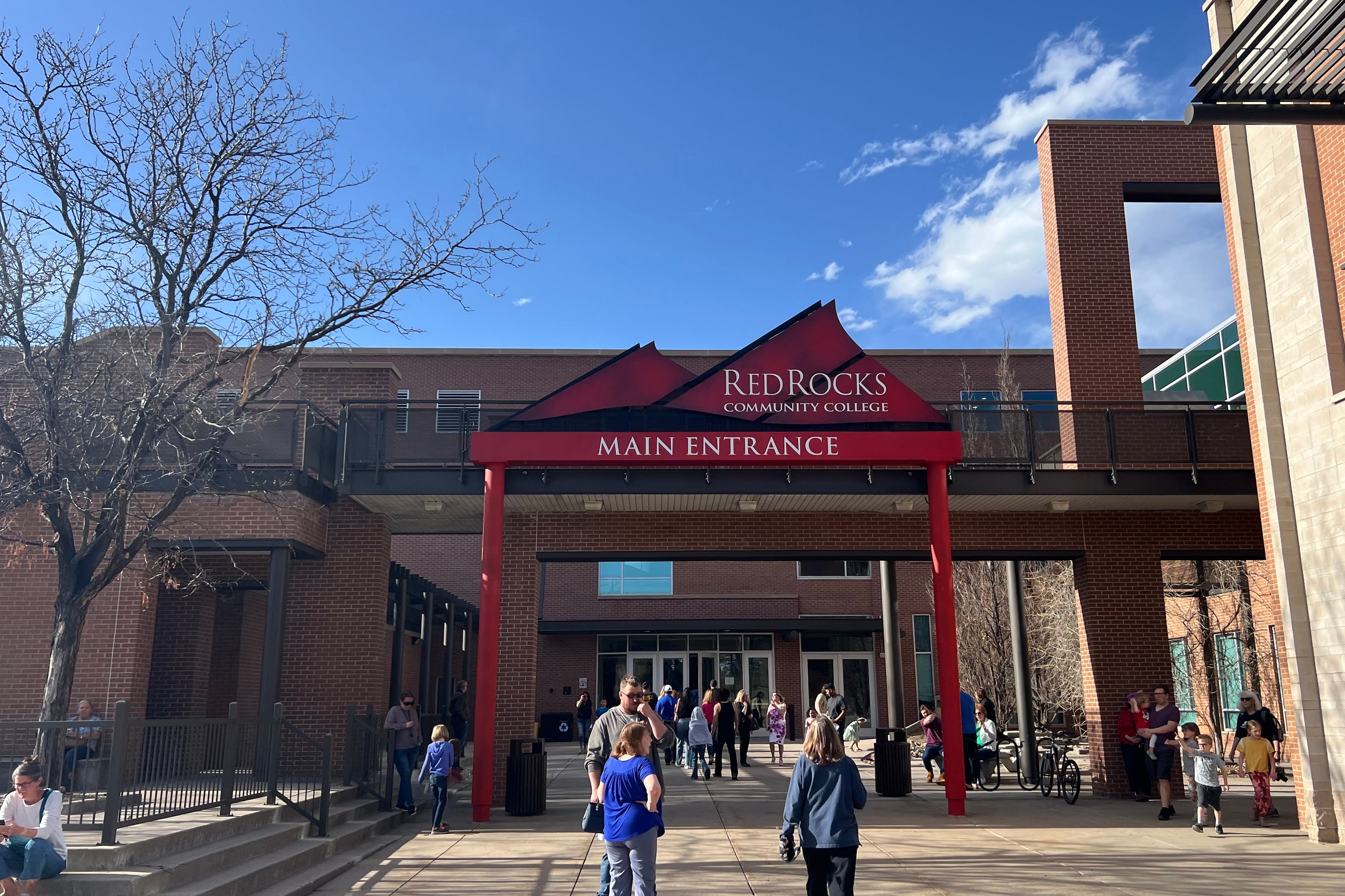 People walk on a concrete sidewalk near large buildings and a red sign that says "Red Rocks Community College" with a blue sky in the background.