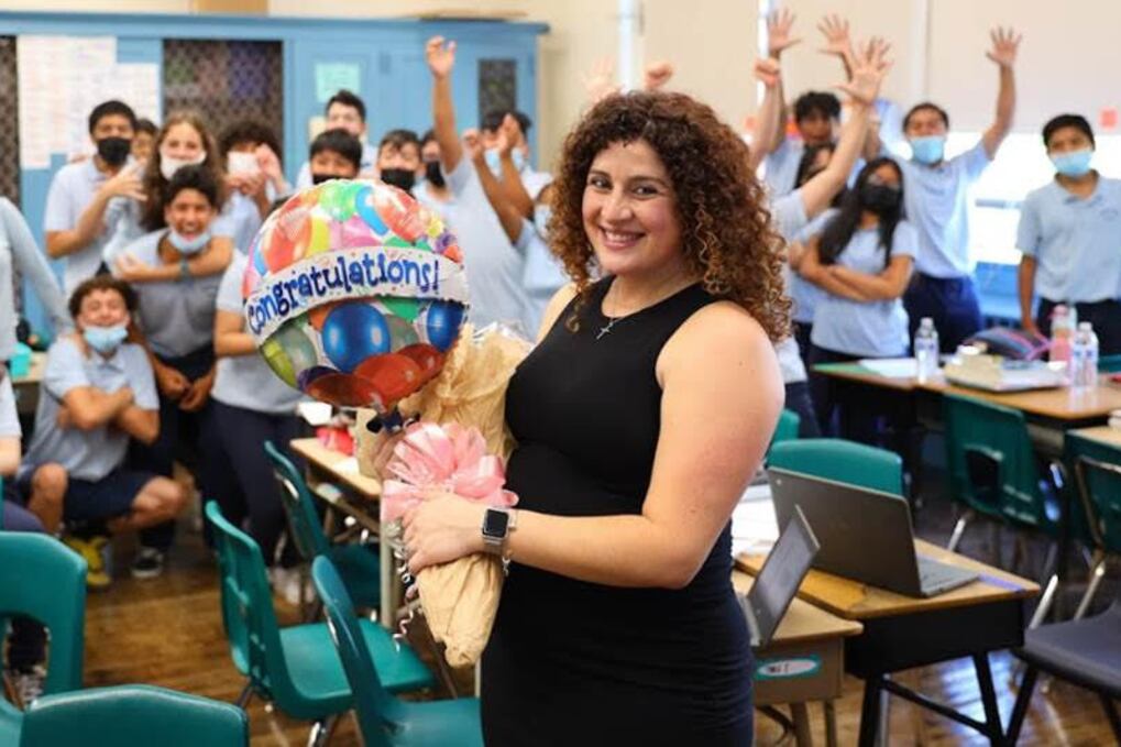 Jessica Tavares in a black dress smiles as she holds a balloon with the word congratulations and her students cheer her on in the background.