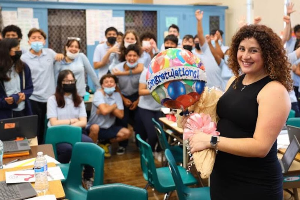 Jessica Tavares in a black dress smiles as she holds a balloon with the word congratulations and her students cheer her on in the background.