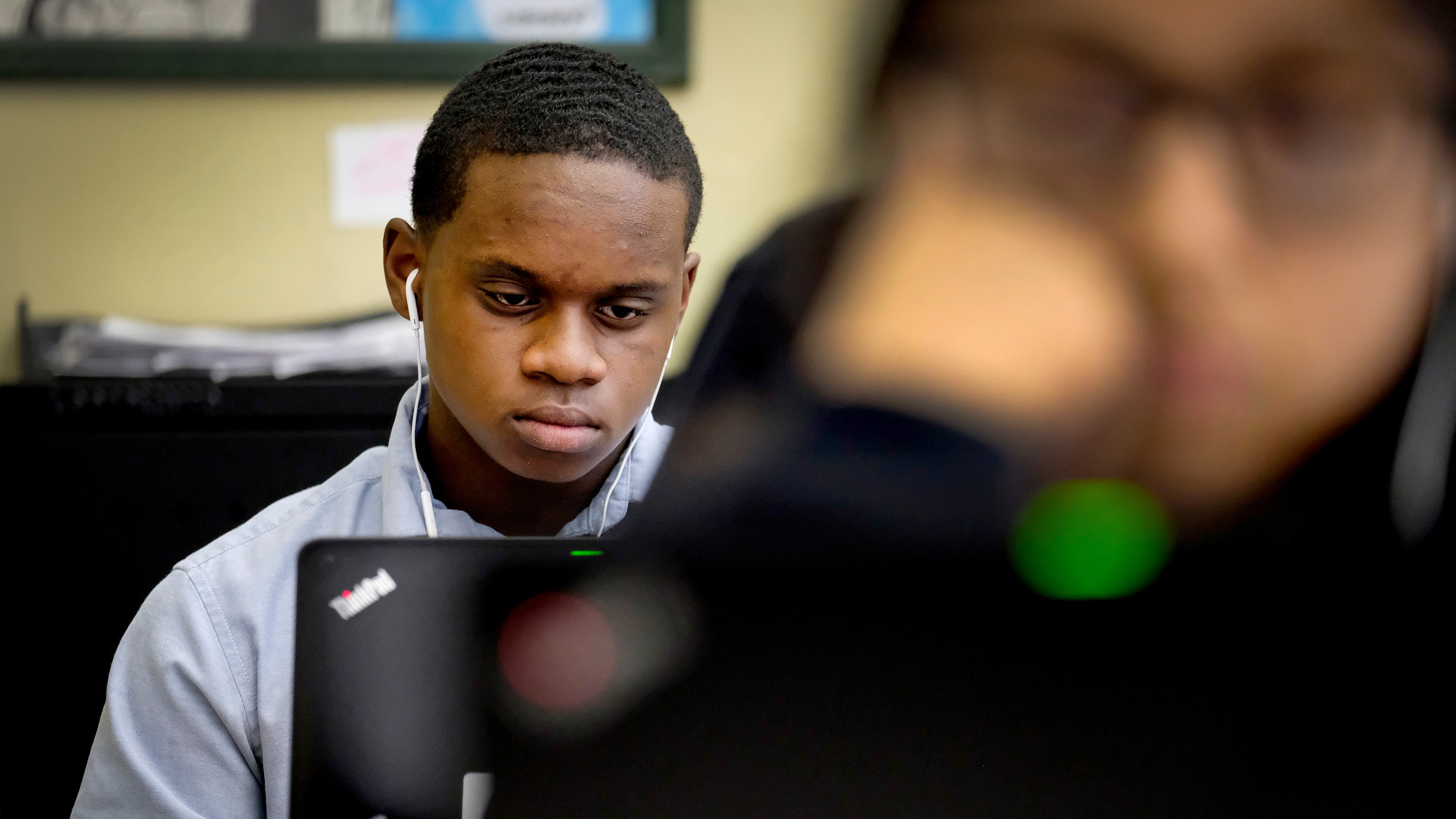 An Indiana high school student uses a laptop computer; in front of him in the foreground is the blurred image of another student staring toward a desk.