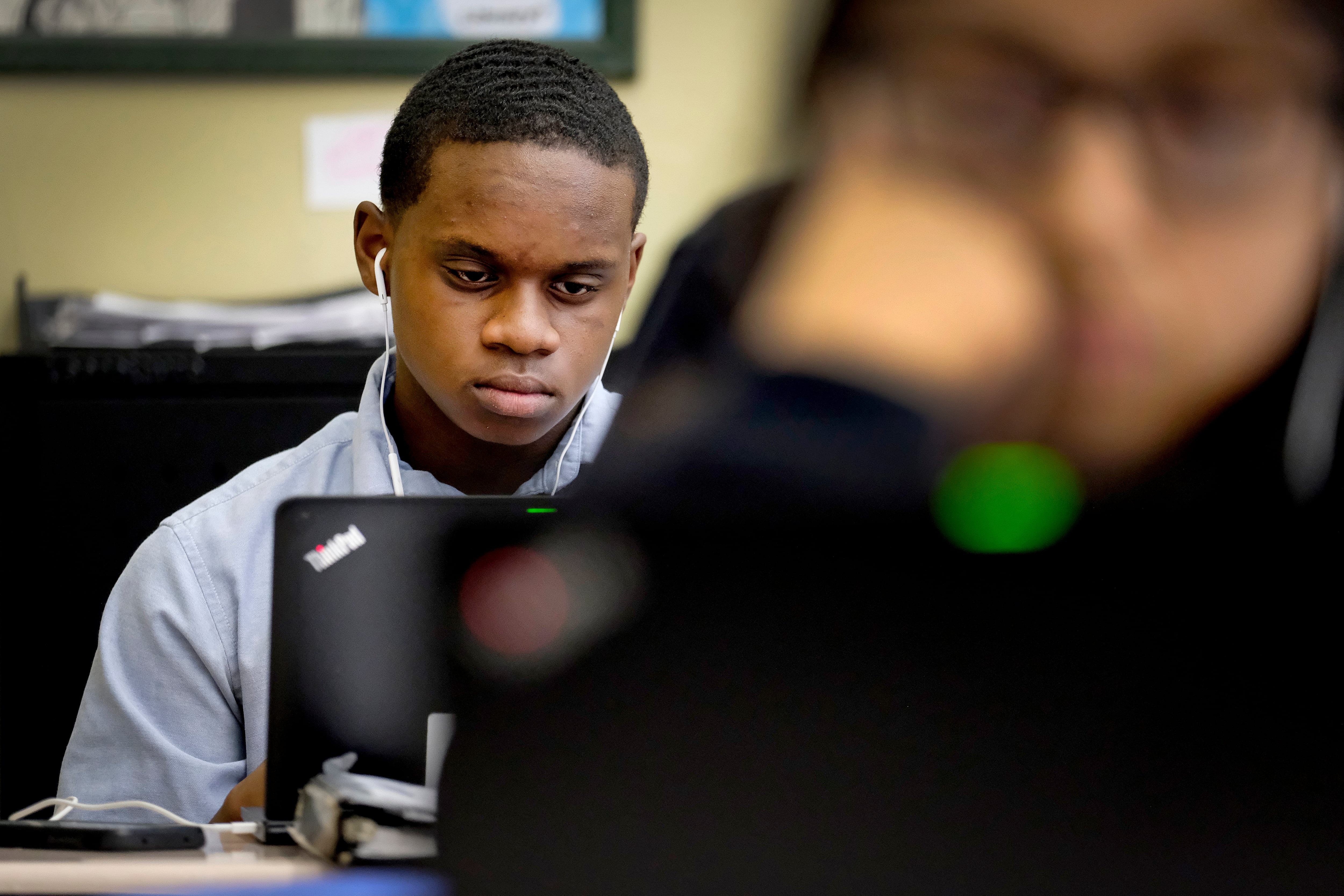 An Indiana high school student uses a laptop computer; in front of him in the foreground is the blurred image of another student staring toward a desk.