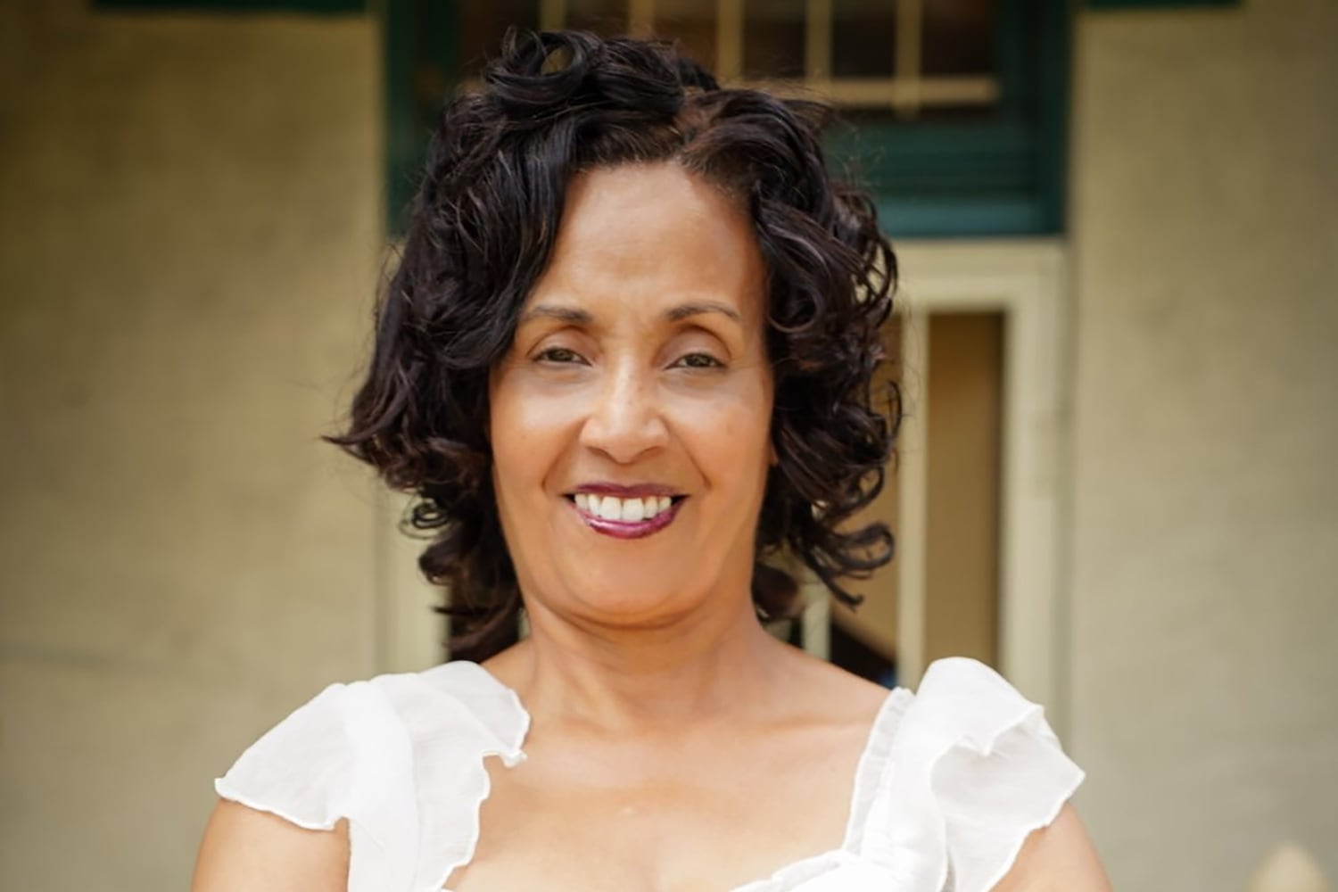 A photograph of a woman with short dark hair and wearing a white blouse poses for a portrait outside.