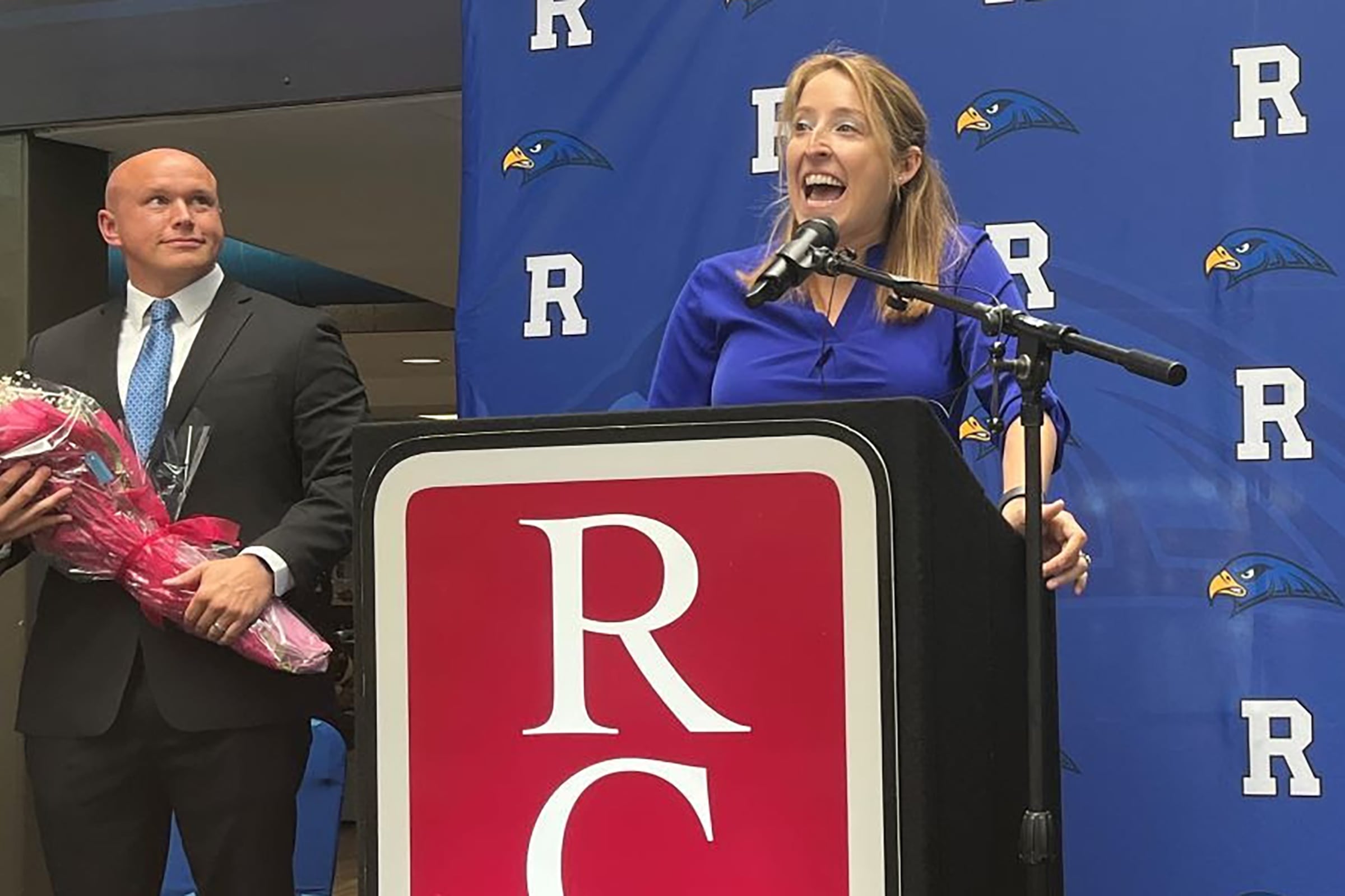 A woman with blonde hair and a blue top speaks at a podium as a man holds flowers behind her.