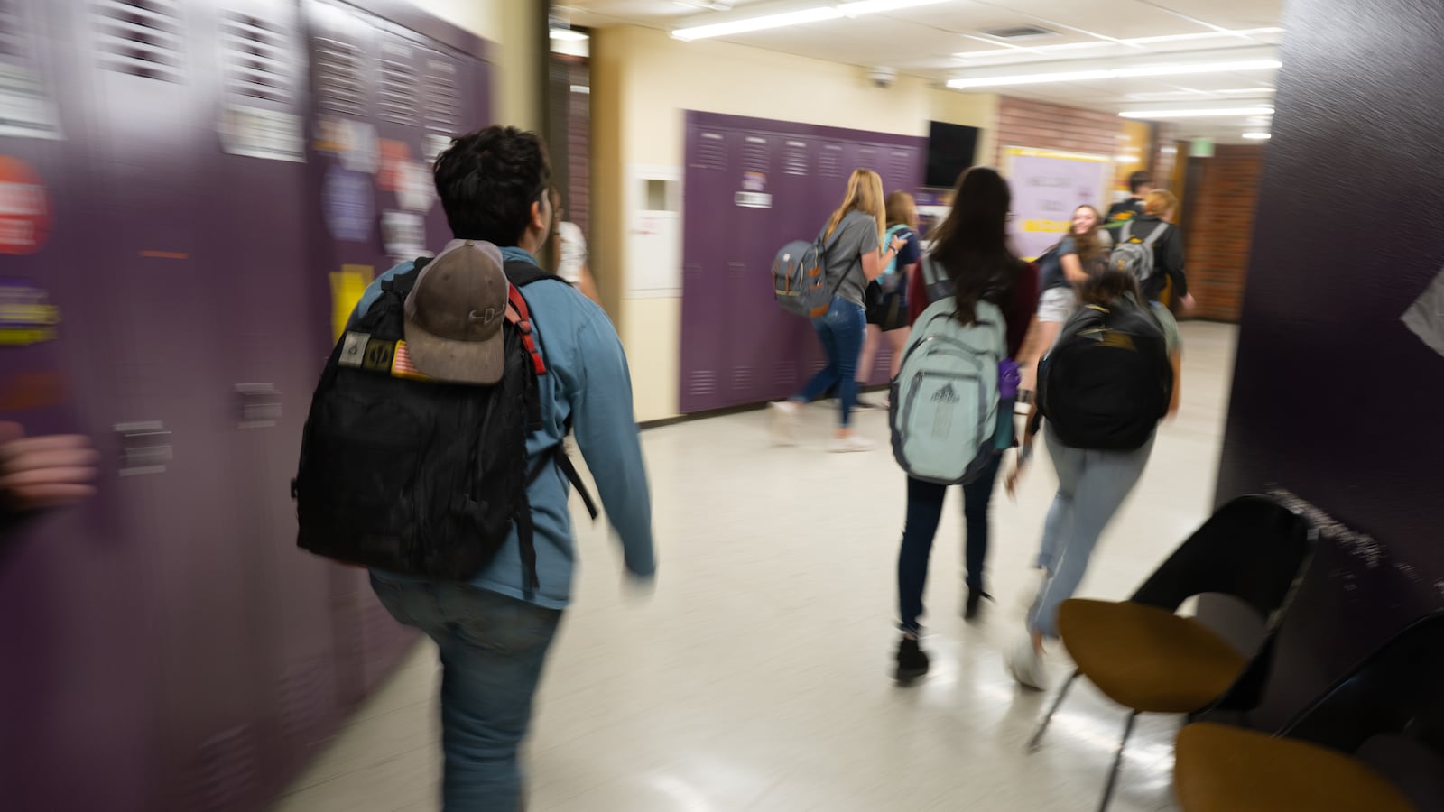 A boy wearing a backpack walks down a school hallway with purple lockers, while a group of students walks ahead of him.