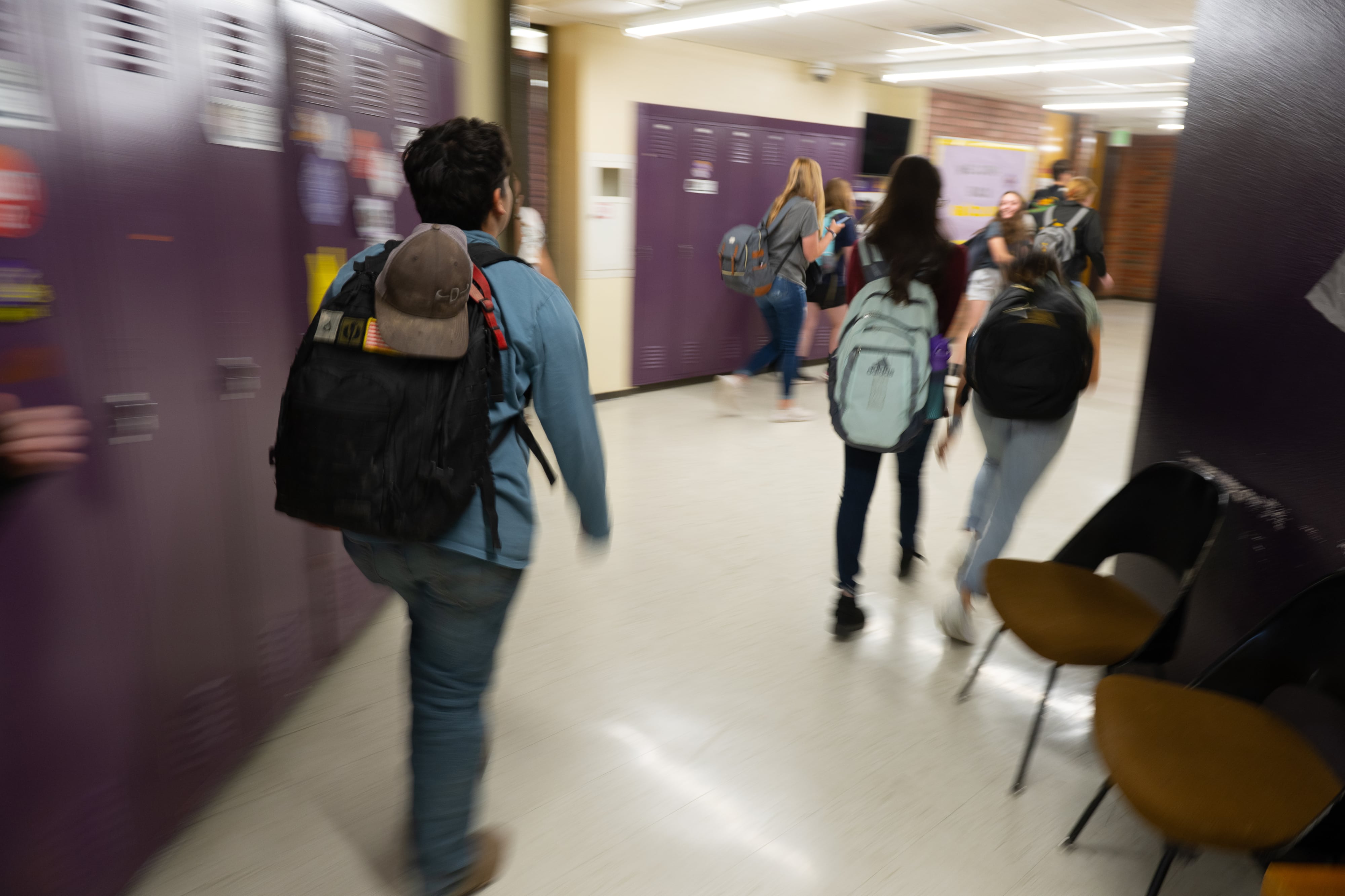 A boy wearing a backpack walks down a school hallway with purple lockers, while a group of students walks ahead of him.