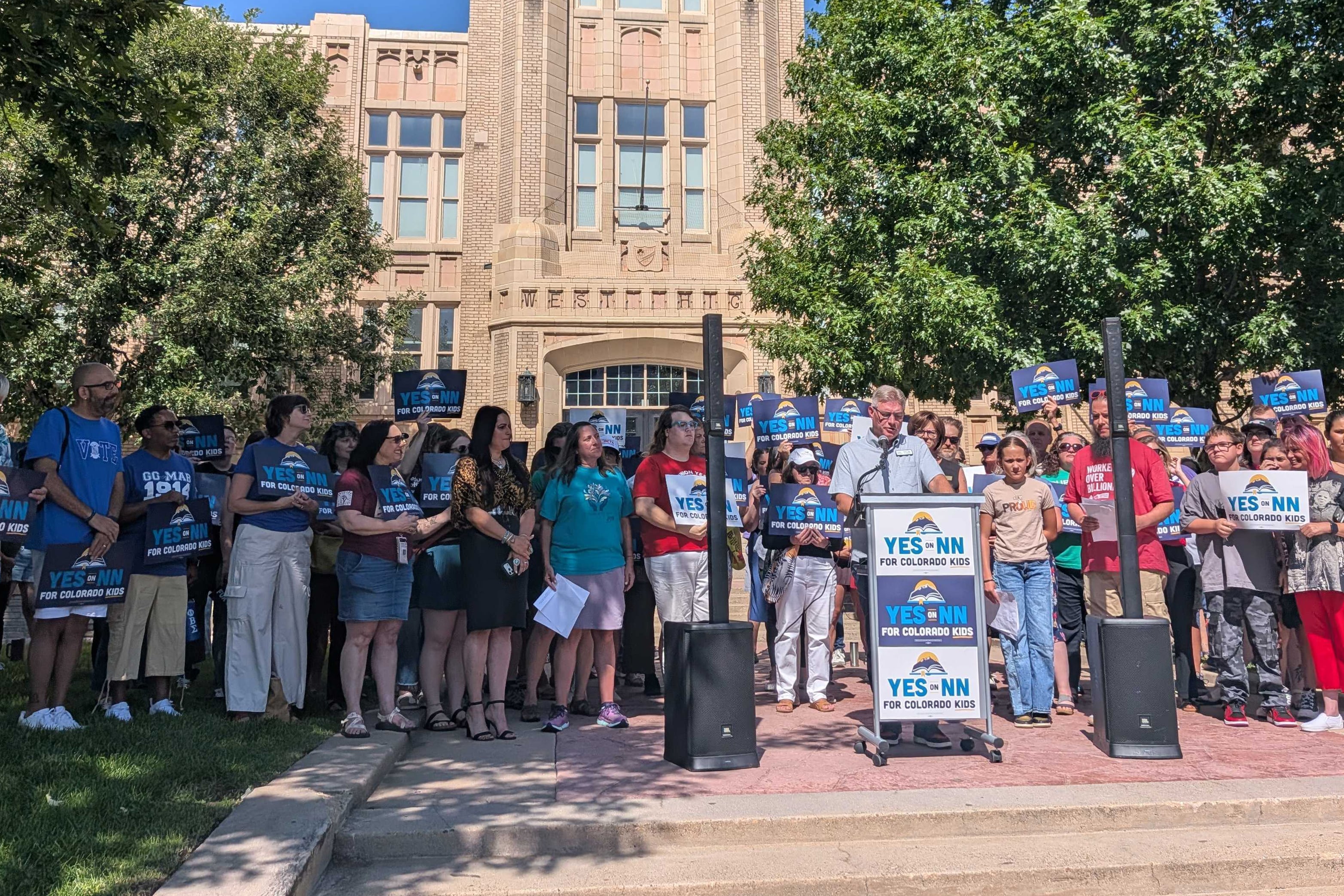 A large group of people stand behind a podium holding signs during a rally with a large stone building in the background.