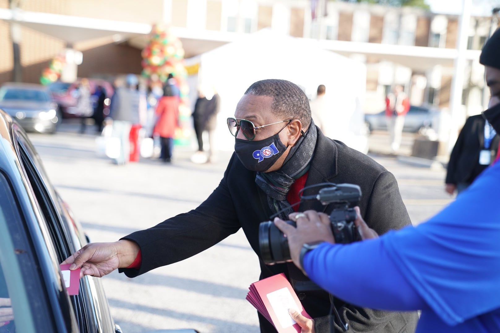 Superintendent Joris Ray hands a gift to a family through a car window as he stands outside the district’s central office in a mask and sunglasses.