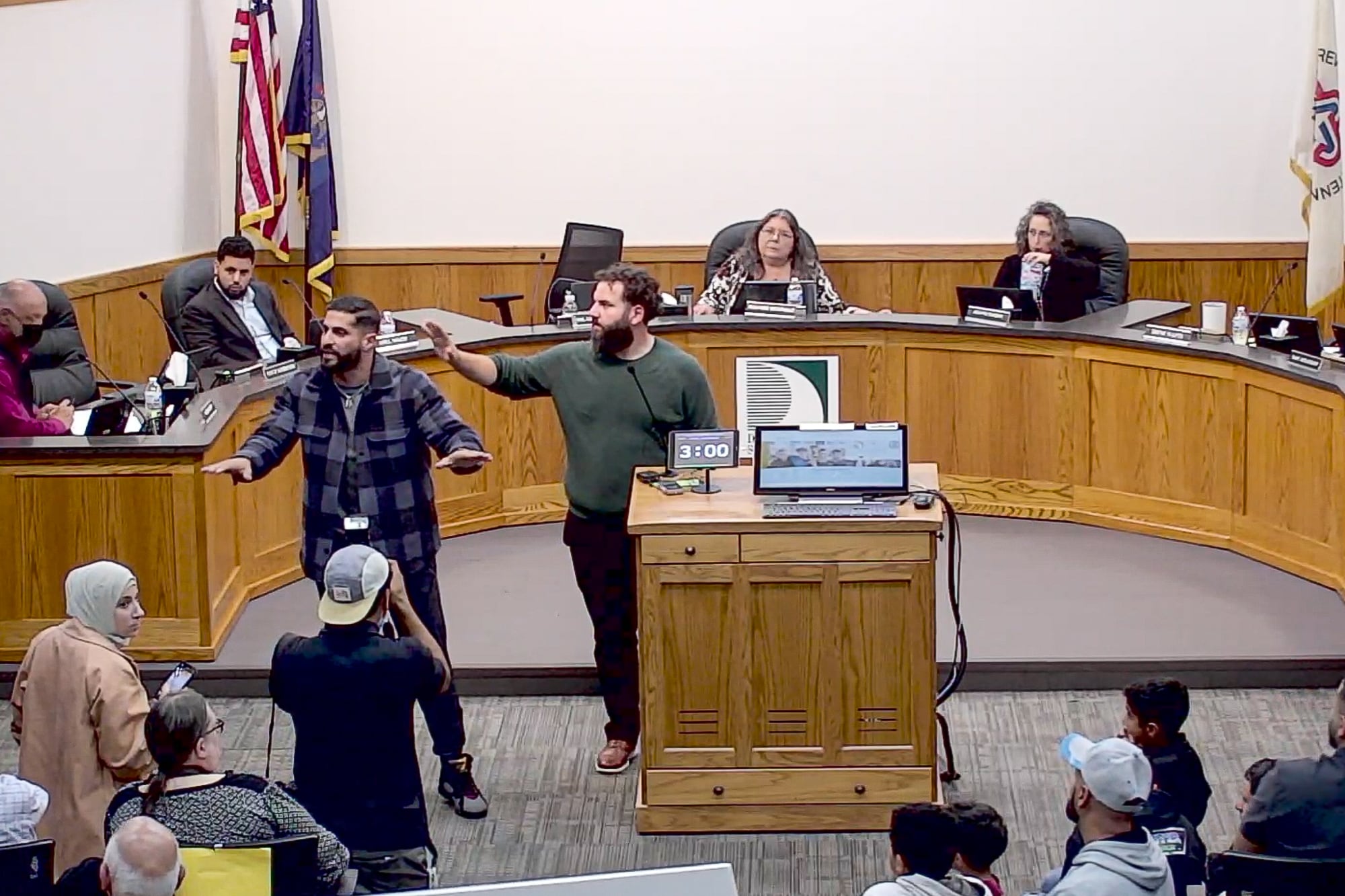 two men stand with arms up trying to calm a crowd in an administrative building as board members sit behind a semicircle desk in the background