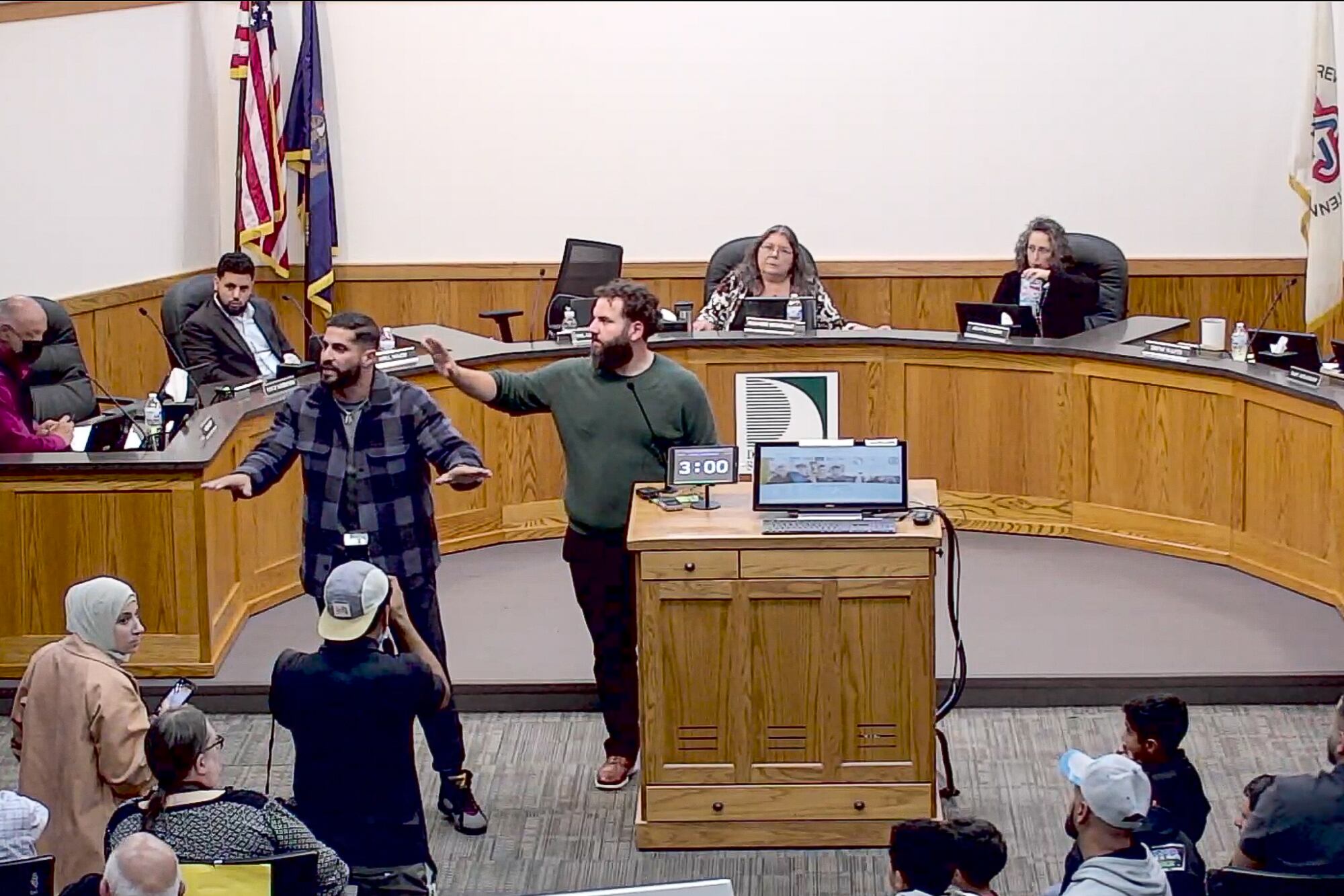 two men stand with arms up trying to calm a crowd in an administrative building as board members sit behind a semicircle desk in the background