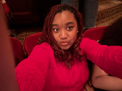 A photograph a Black high school student with short dark hair and wearing a pink sweater poses for a selfie.