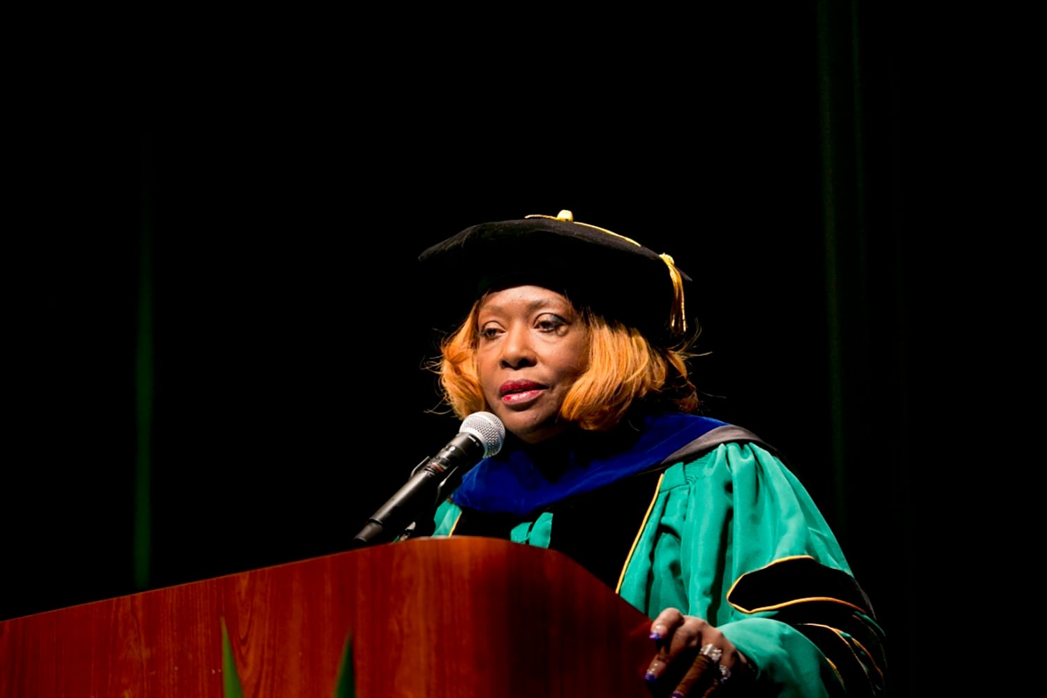 A Black woman wearing Doctorate graduation regalia speaks from a wooden podium during a graduation ceremony.