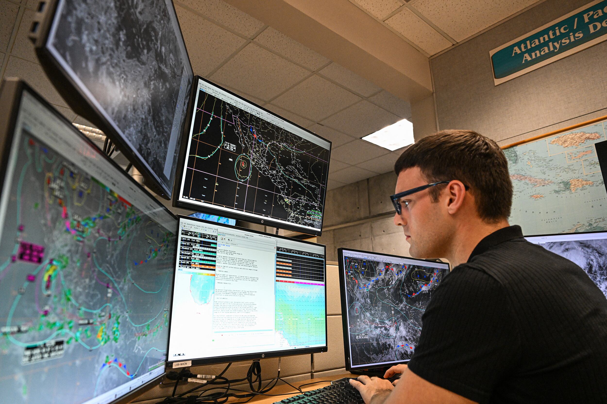 A photograph of a white man with short brown hair, wearing glasses and a black t-shirt looking at five different large computer screens in an office.