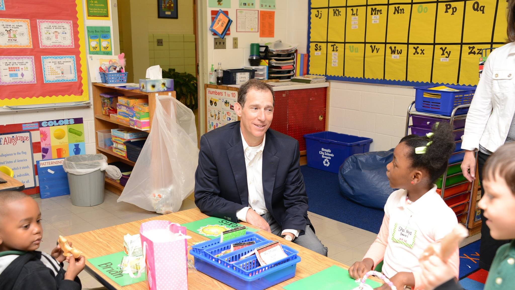 A male politician in a suit jacket sits at a small table in an elementary school classroom with two young students. A young girl looks at him inquisitively.