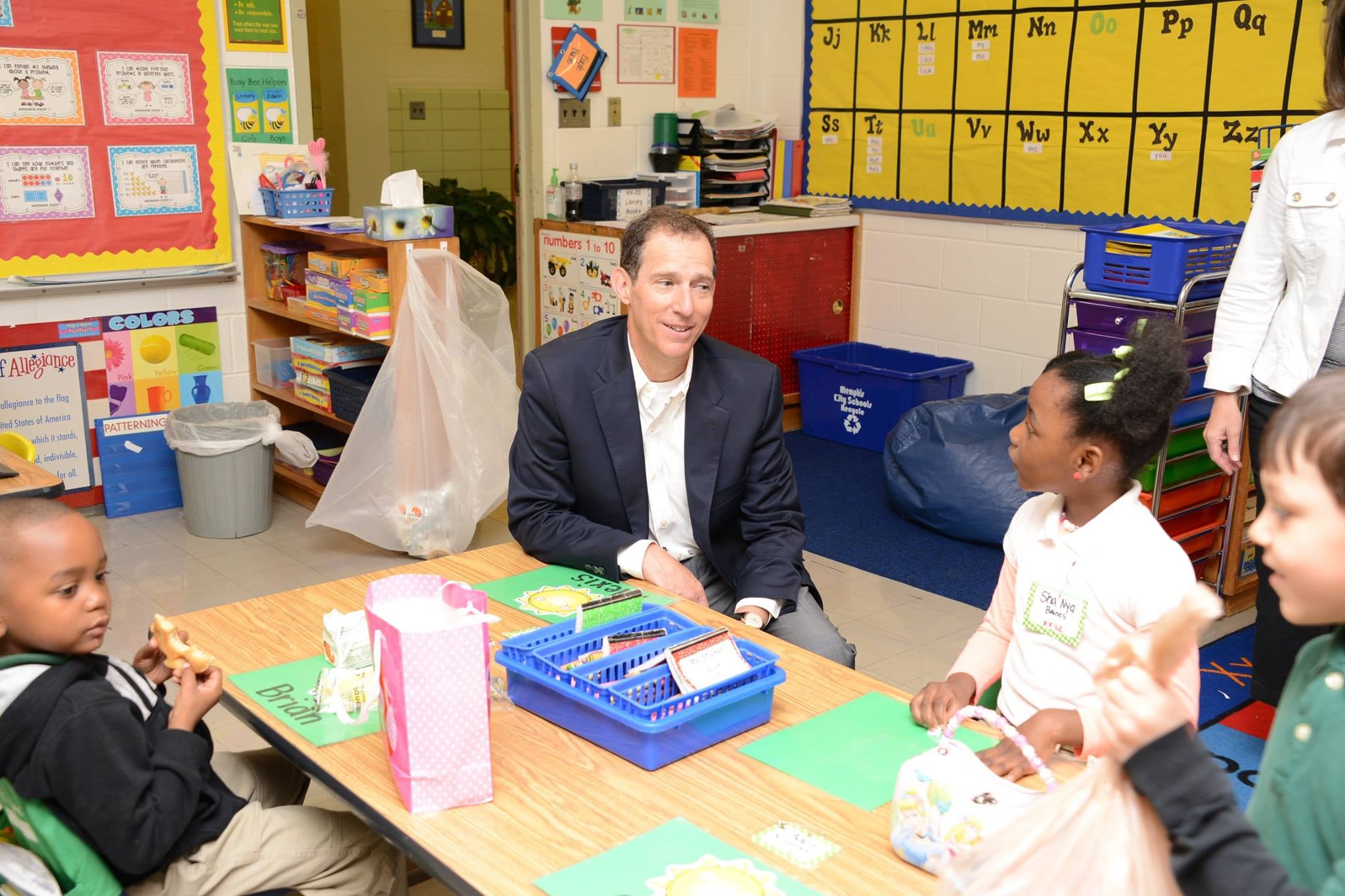 A male politician in a suit jacket sits at a small table in an elementary school classroom with two young students. A young girl looks at him inquisitively.