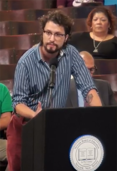 A screen grab of a board meeting with a man standing at a microphone during public comment.