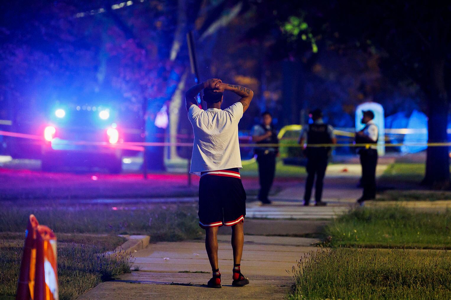A person stands with their back toward the camera and their hands behind their head while they look at police behind caution tape.