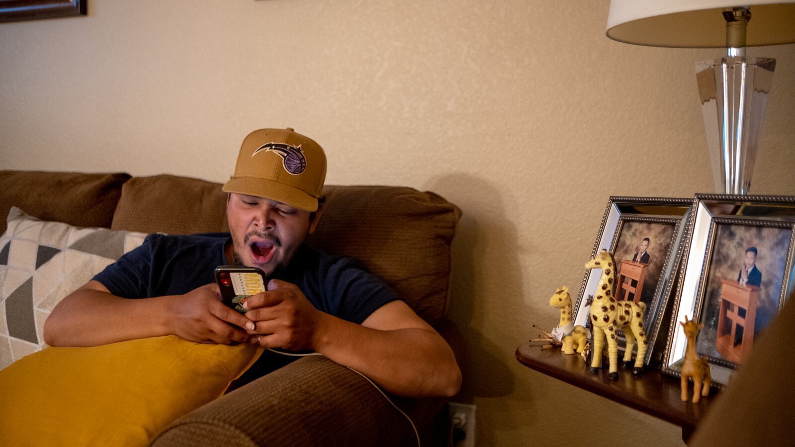A young man, wearing a tan Orlando Magic hat, yawns as he looks at his phone while sitting on the couch.