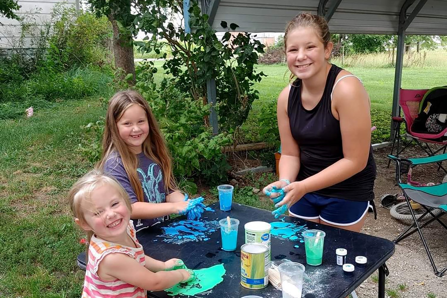 Three young children stand around a table outside smiling at the camera.
