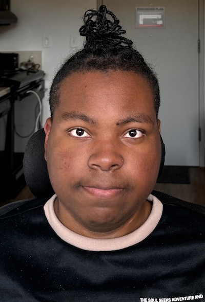 A high school boy wearing a black shirt and has his hair tied up on his head poses for a portrait inside of a room.