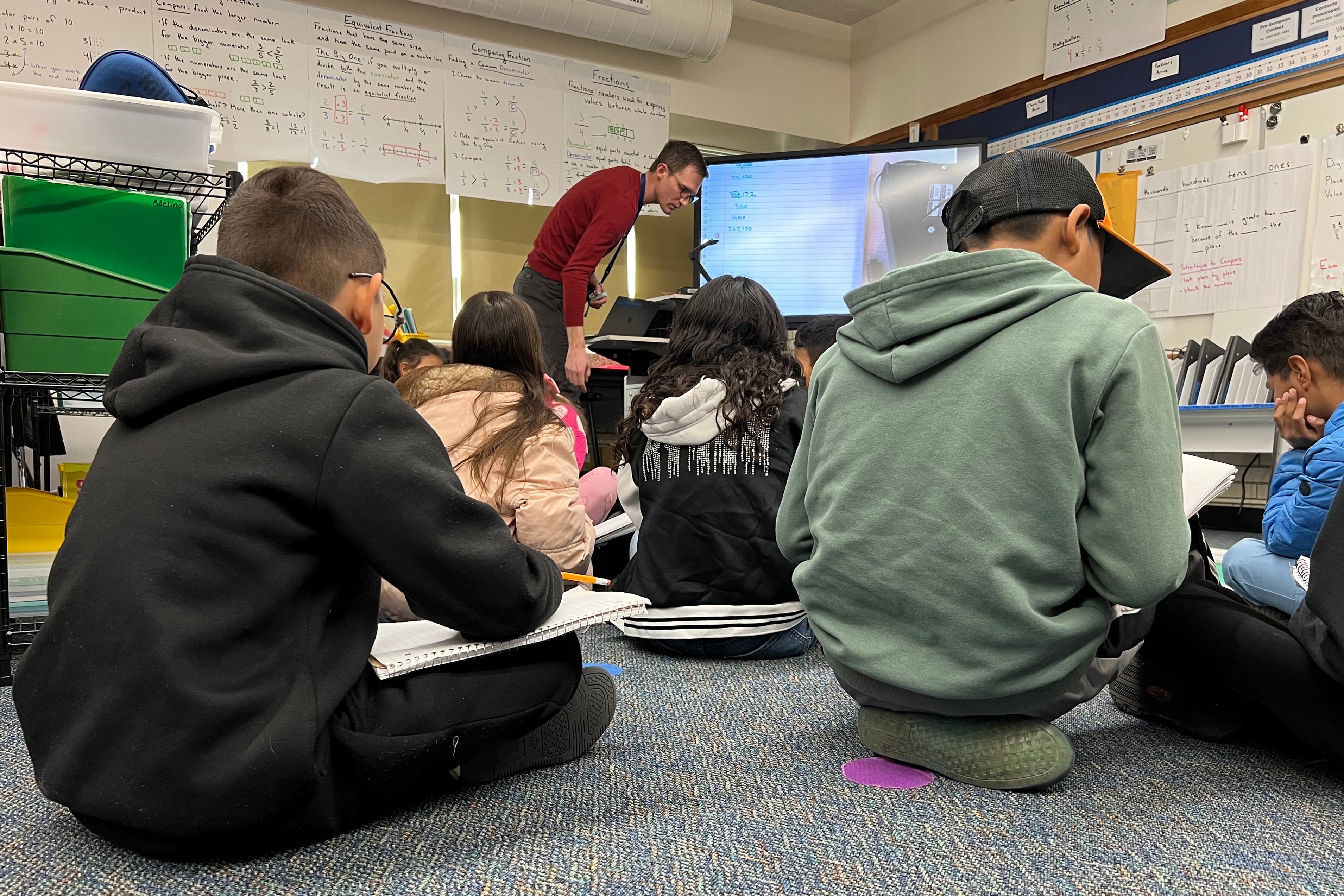 A group of elementary students sit on grey carpet while a teacher in a red shirt stands at the front of the classroom.