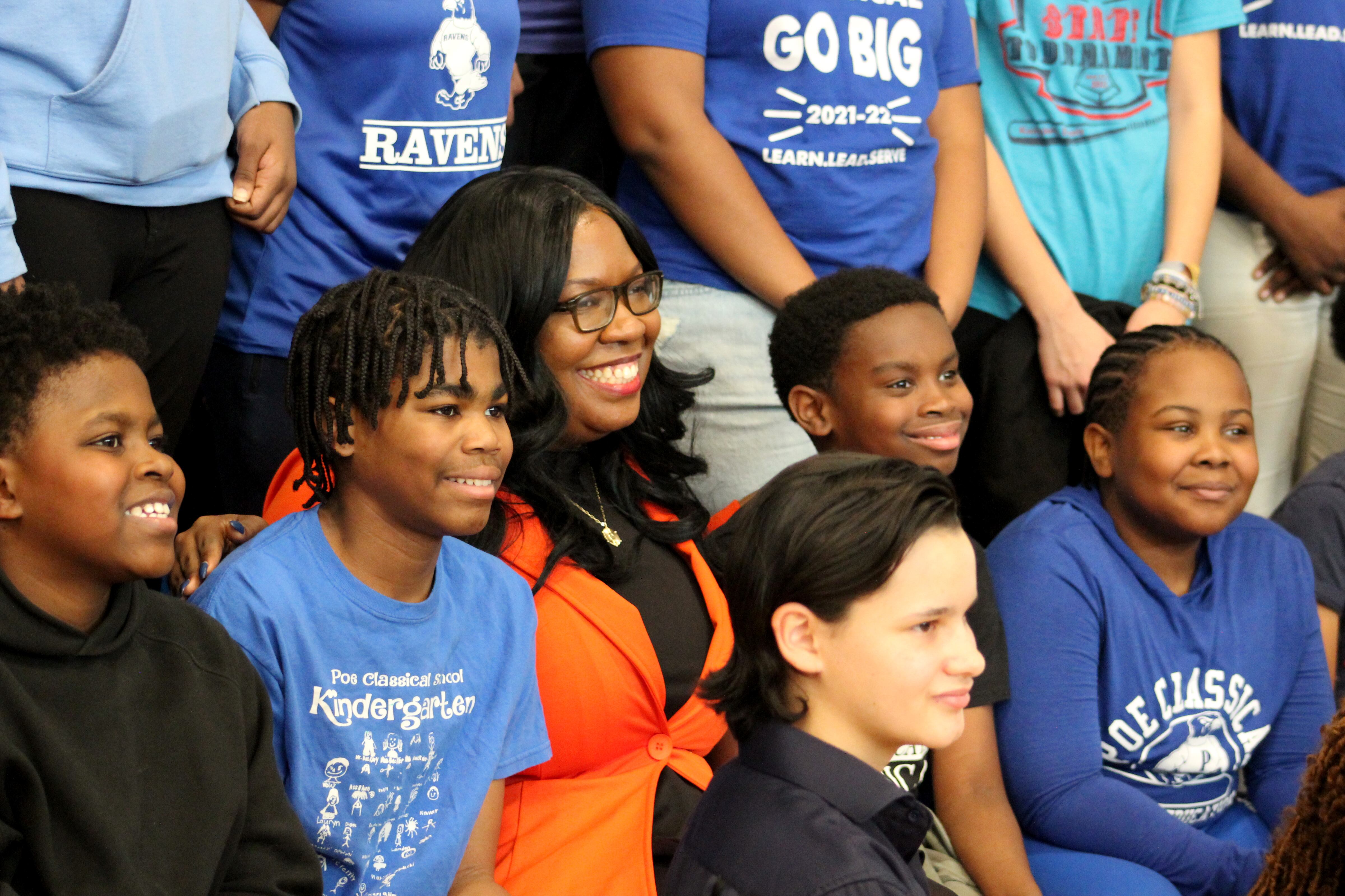An adult female with long black hair and wearing an orange suit jacket sits between a crowd of students smiling for the camera.