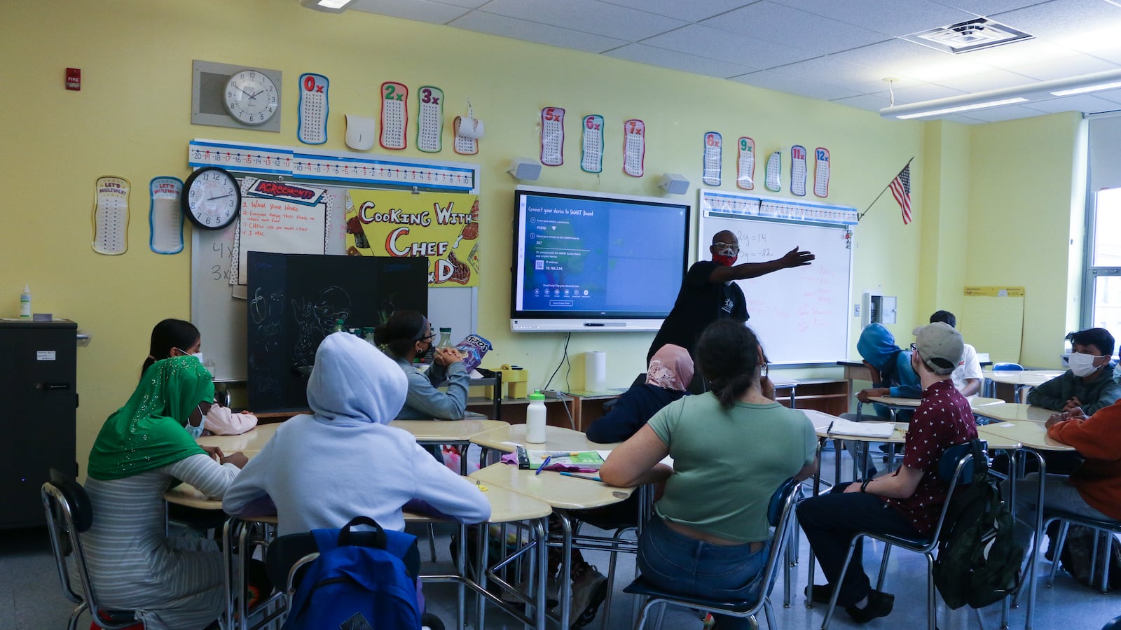 Students sit in a classroom wearing masks while a teacher leads a lesson in front of a screen.