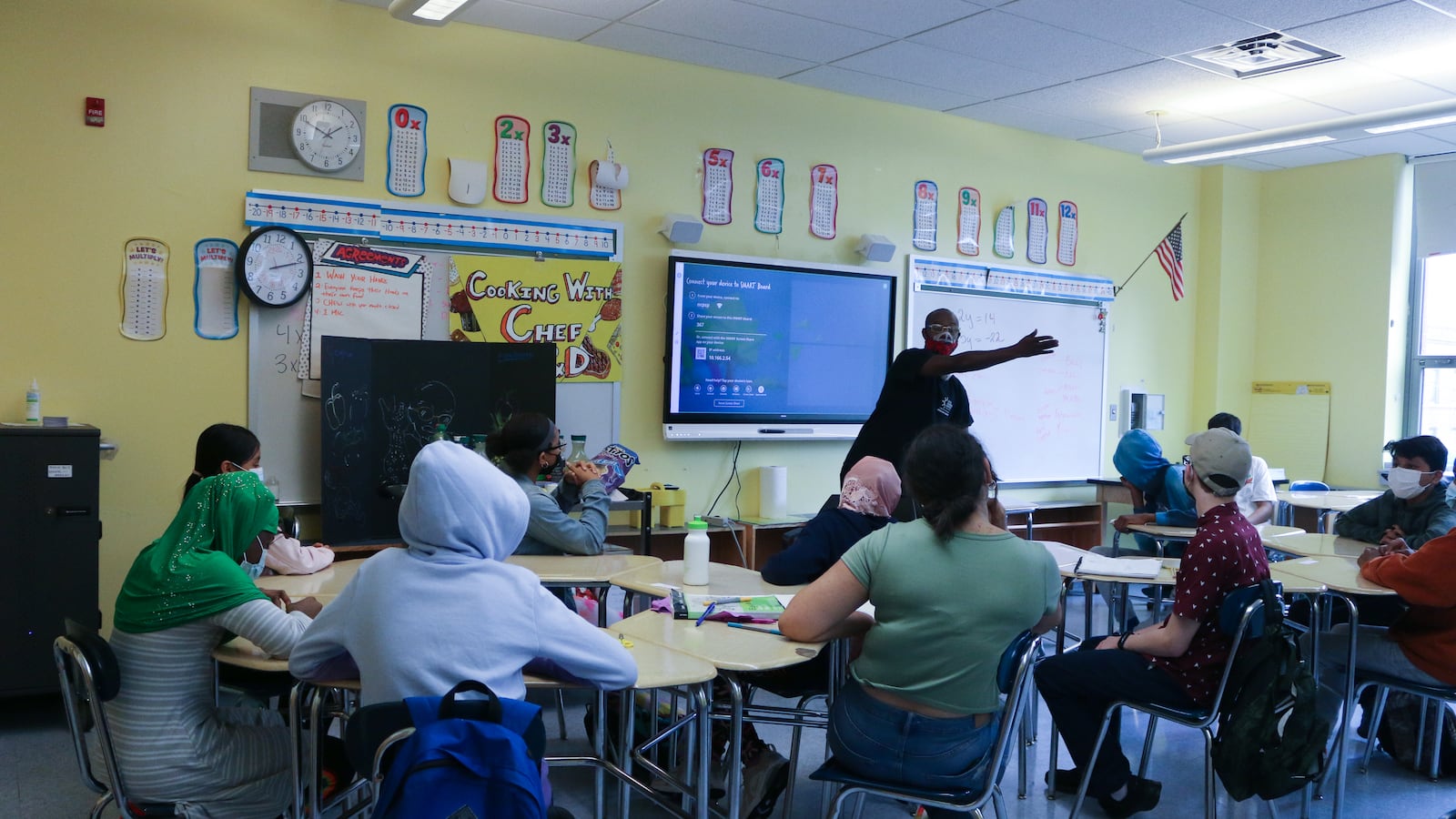 Students sit in a classroom wearing masks while a teacher leads a lesson in front of a screen.