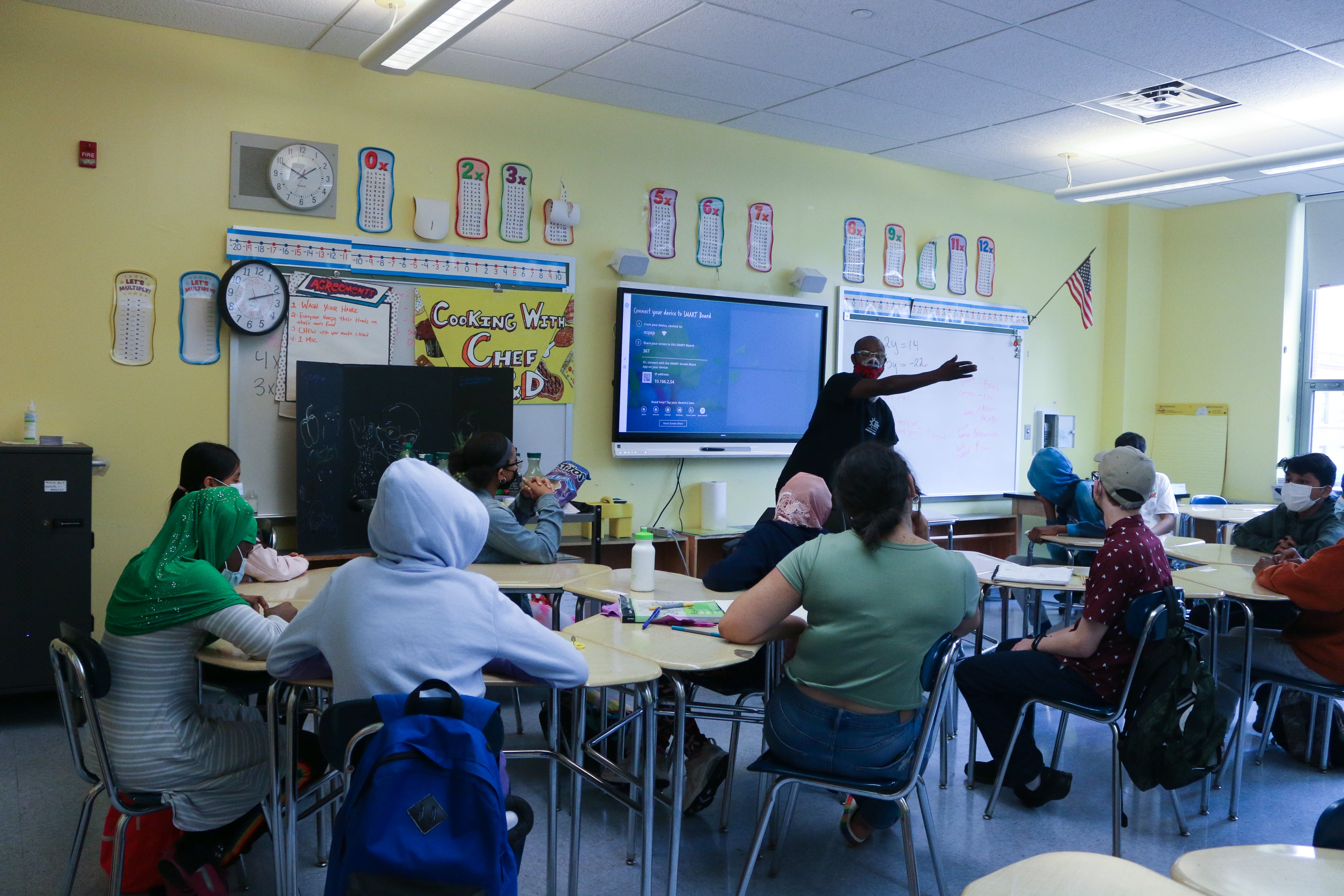 Students sit in a classroom wearing masks while a teacher leads a lesson in front of a screen.