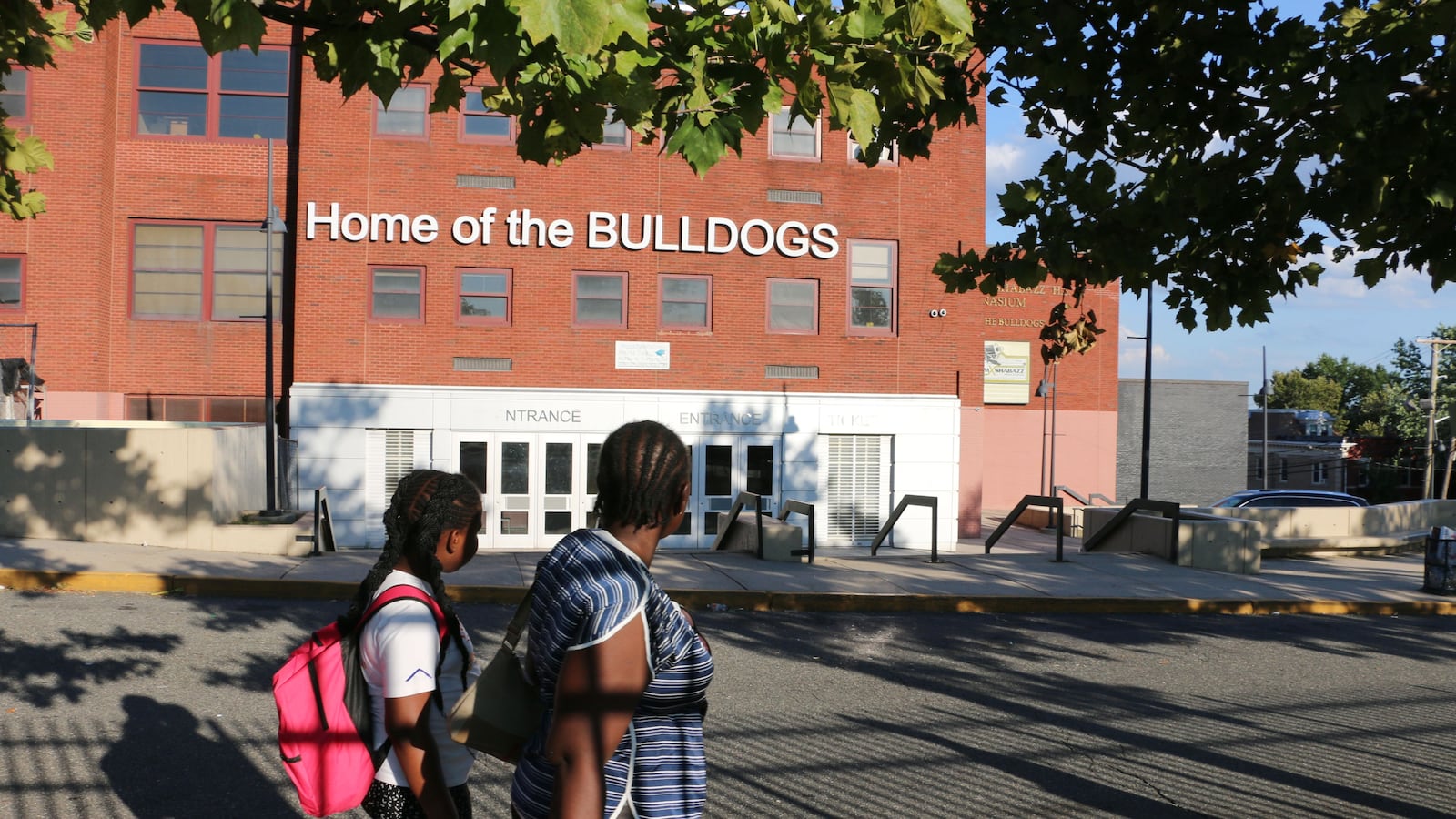 In the foreground, a student wearing a pink backpack stands behind an adult in a blue-striped shirt as they face Malcolm X Shabazz High School in the background.