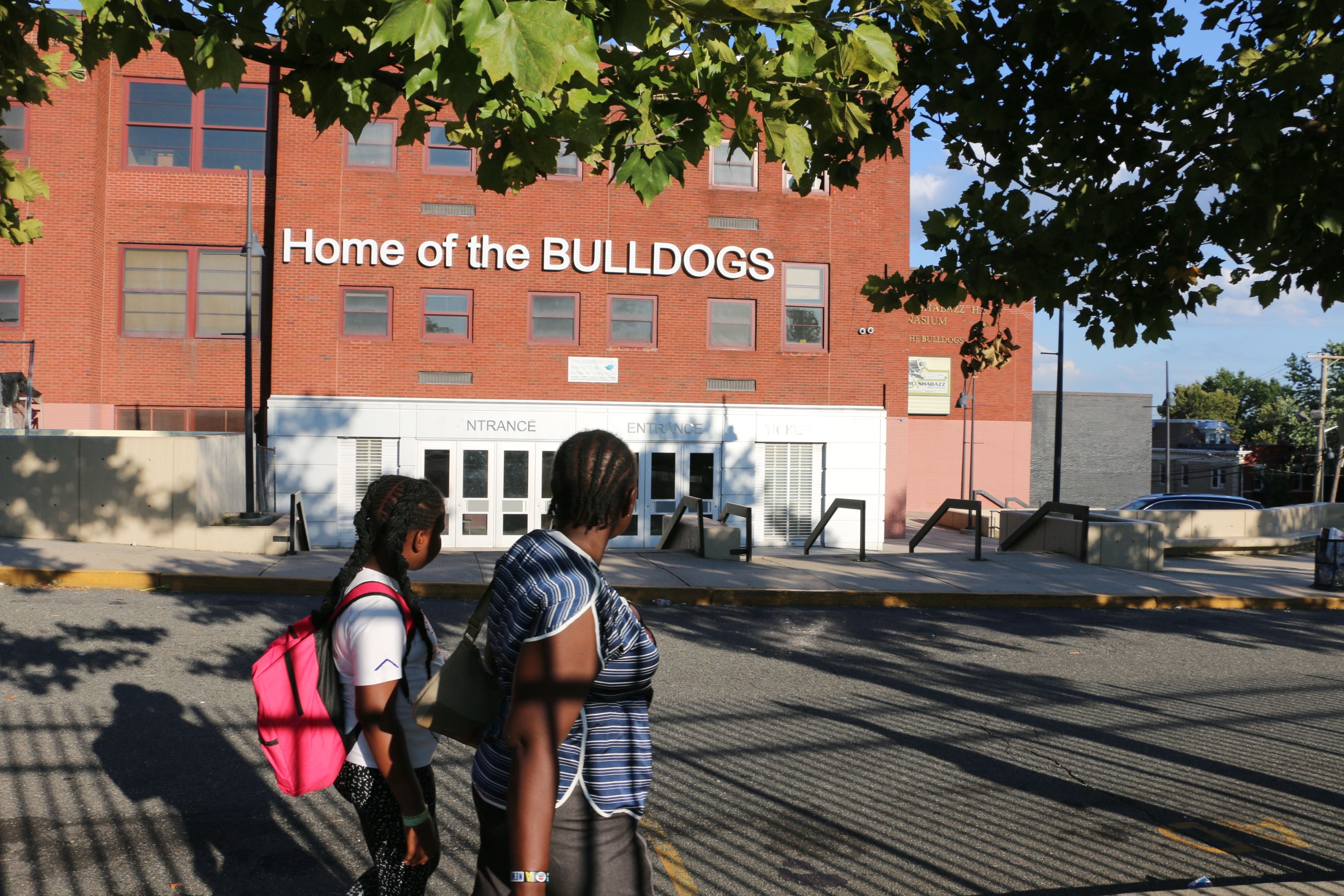 In the foreground, a student wearing a pink backpack stands behind an adult in a blue-striped shirt as they face Malcolm X Shabazz High School in the background.