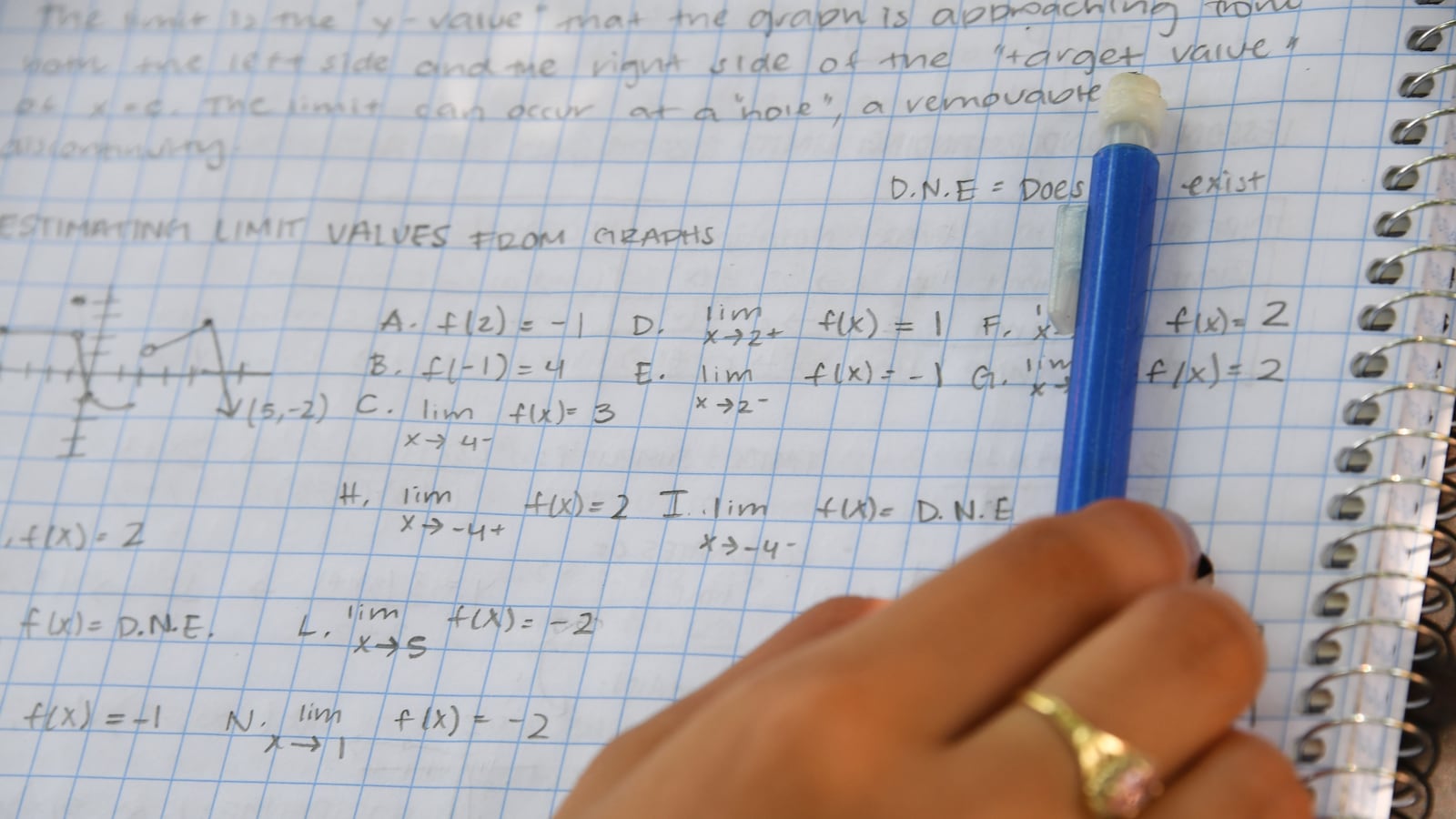 A teenager’s hand holds a blue pencil to a sheet of graph paper, where mathematical equations are written. 