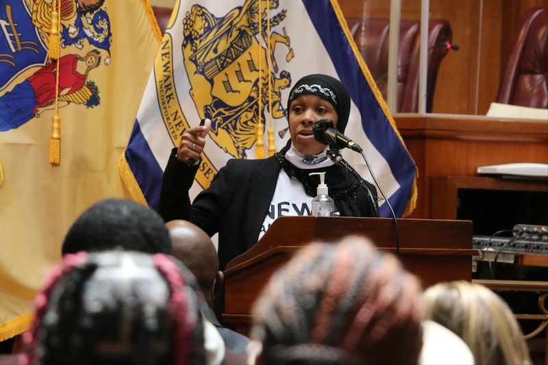 A woman stands at a podium and speaks into a microphone while standing in front of two flags as a crowd watches her.