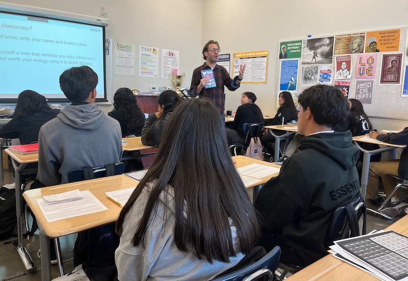 A teacher holds a book and stands at the front of a classroom full of students sitting at desks.