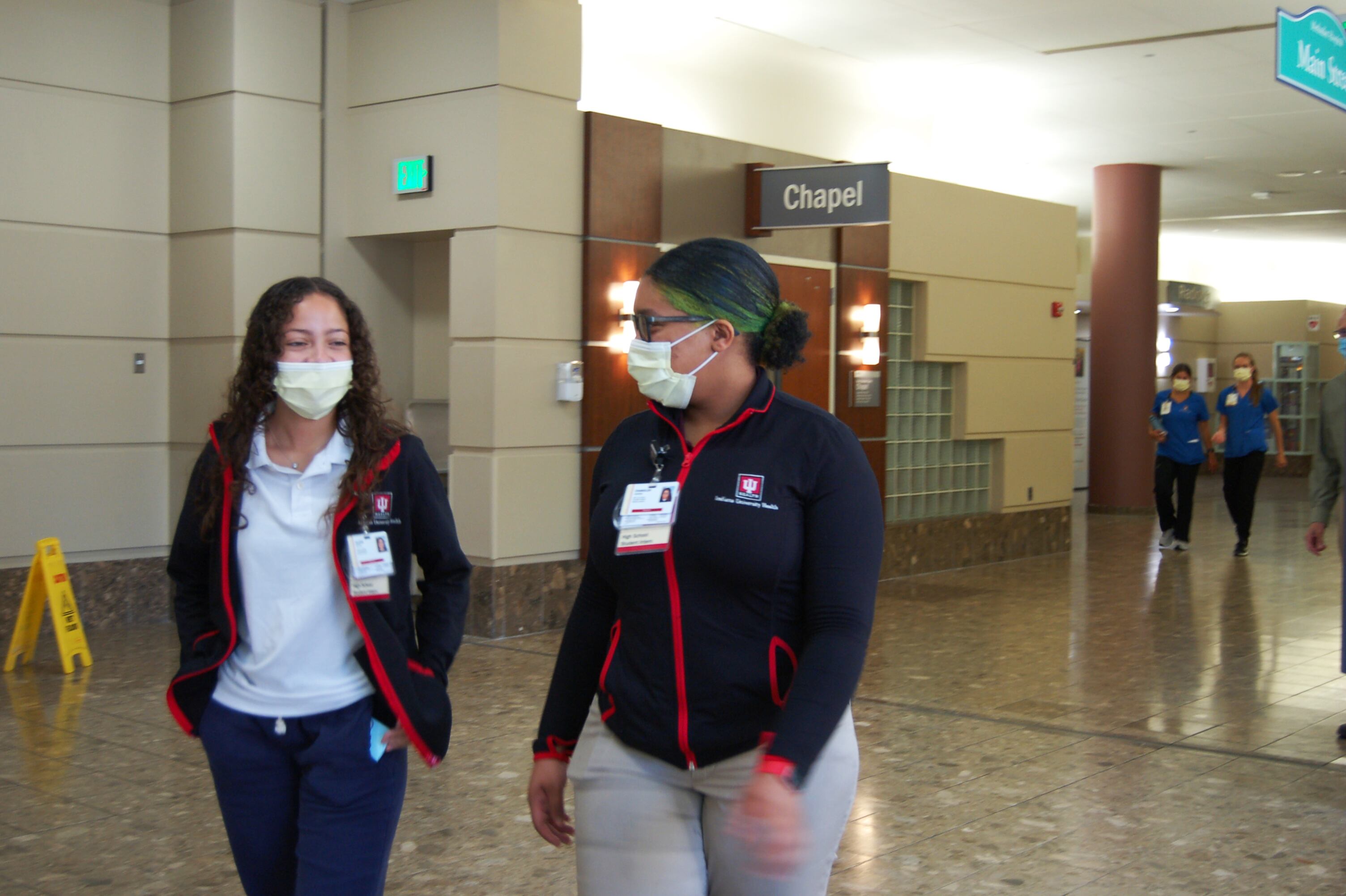 Two girls wearing masks and matching jackets walk down a hospital hallway ahead of health care workers.