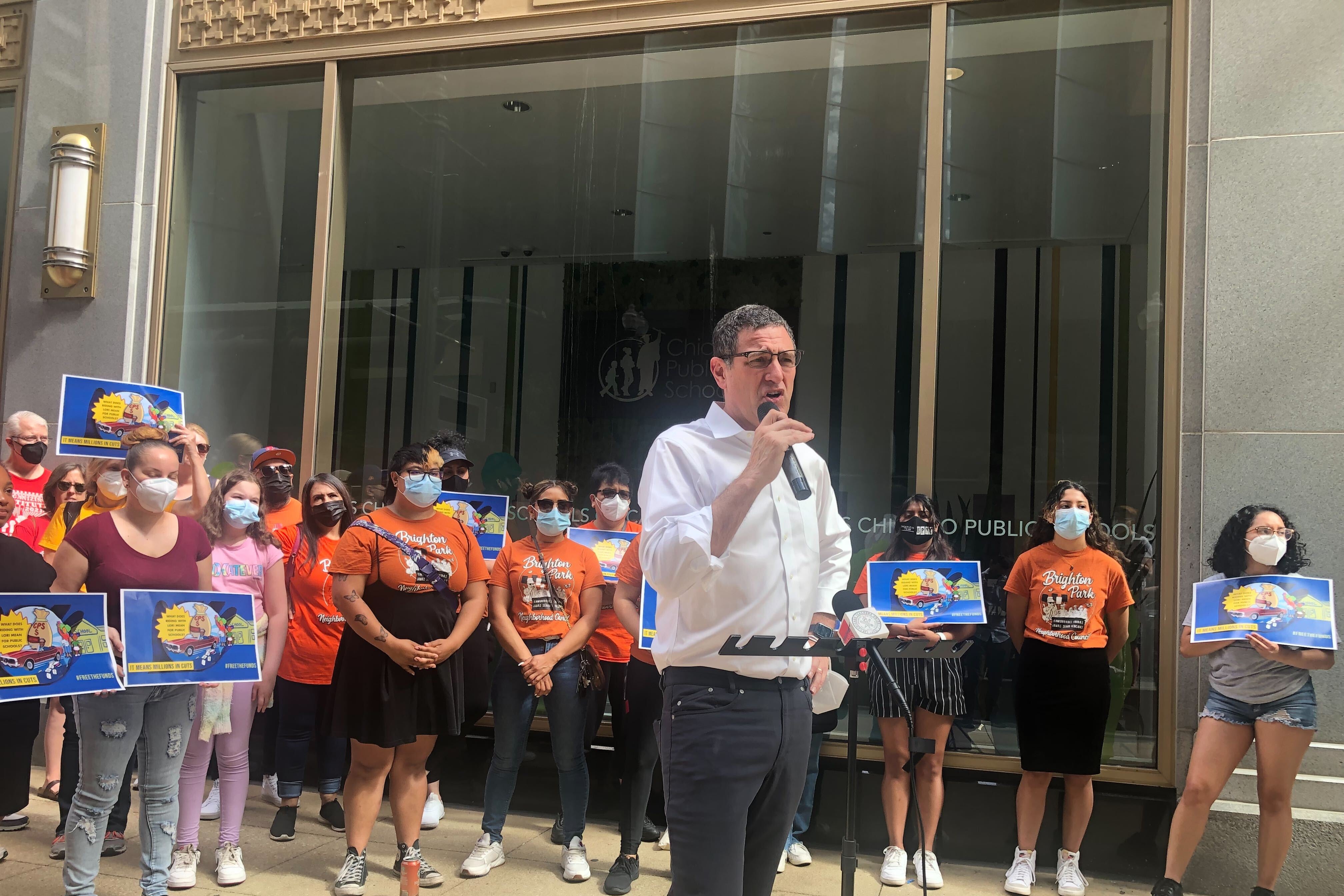 A man in a white shirt, holding a microphone stands in front of a crowd of people outside of Chicago Public School headquarters in downtown.
