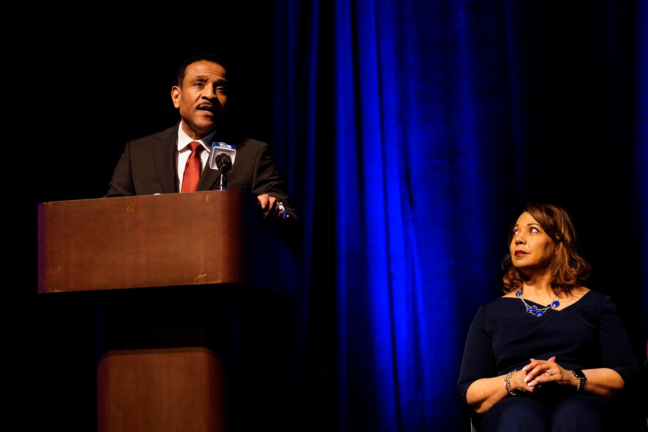 A man wearing a dark suit and a red tie stands at a wooden podium with a dark blue background and a woman sitting on the right.