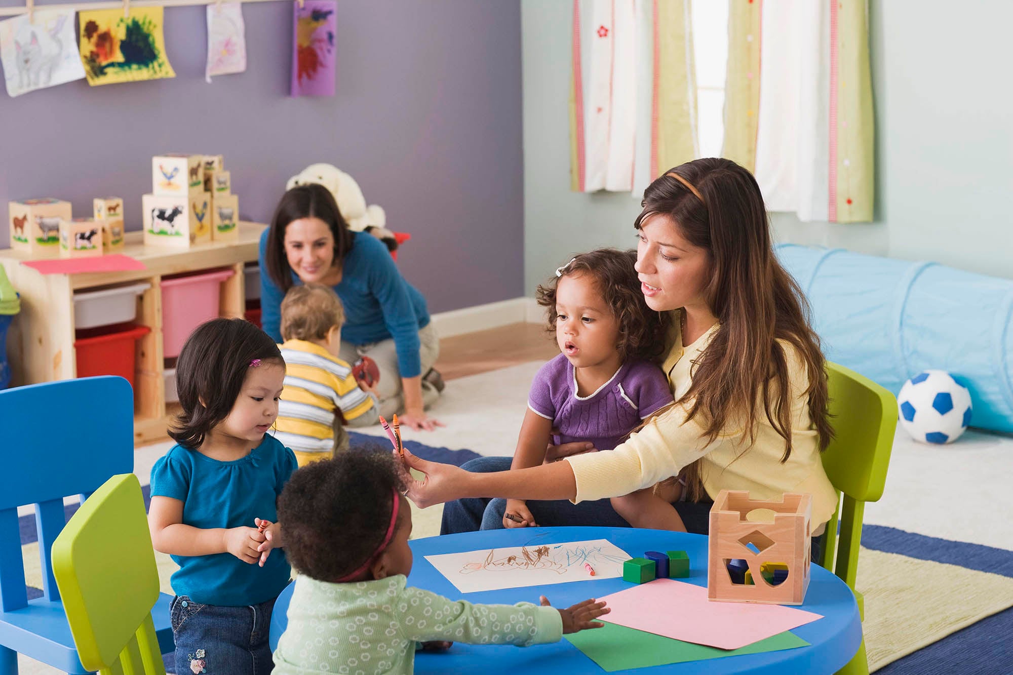Two adults care for four children at a at-home childcare and early education center. There are toys and learning tools in the background with colorful walls.