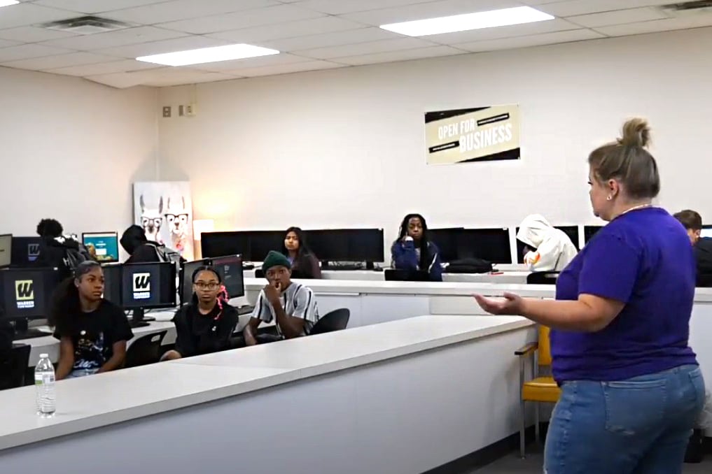 A teacher wearing a purple shirt and jeans stands in front of a high school classroom.