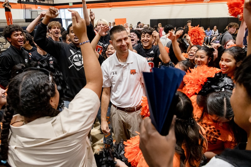 A photograph of a white man in a white shirt stands in front of a large group of students in a gym.