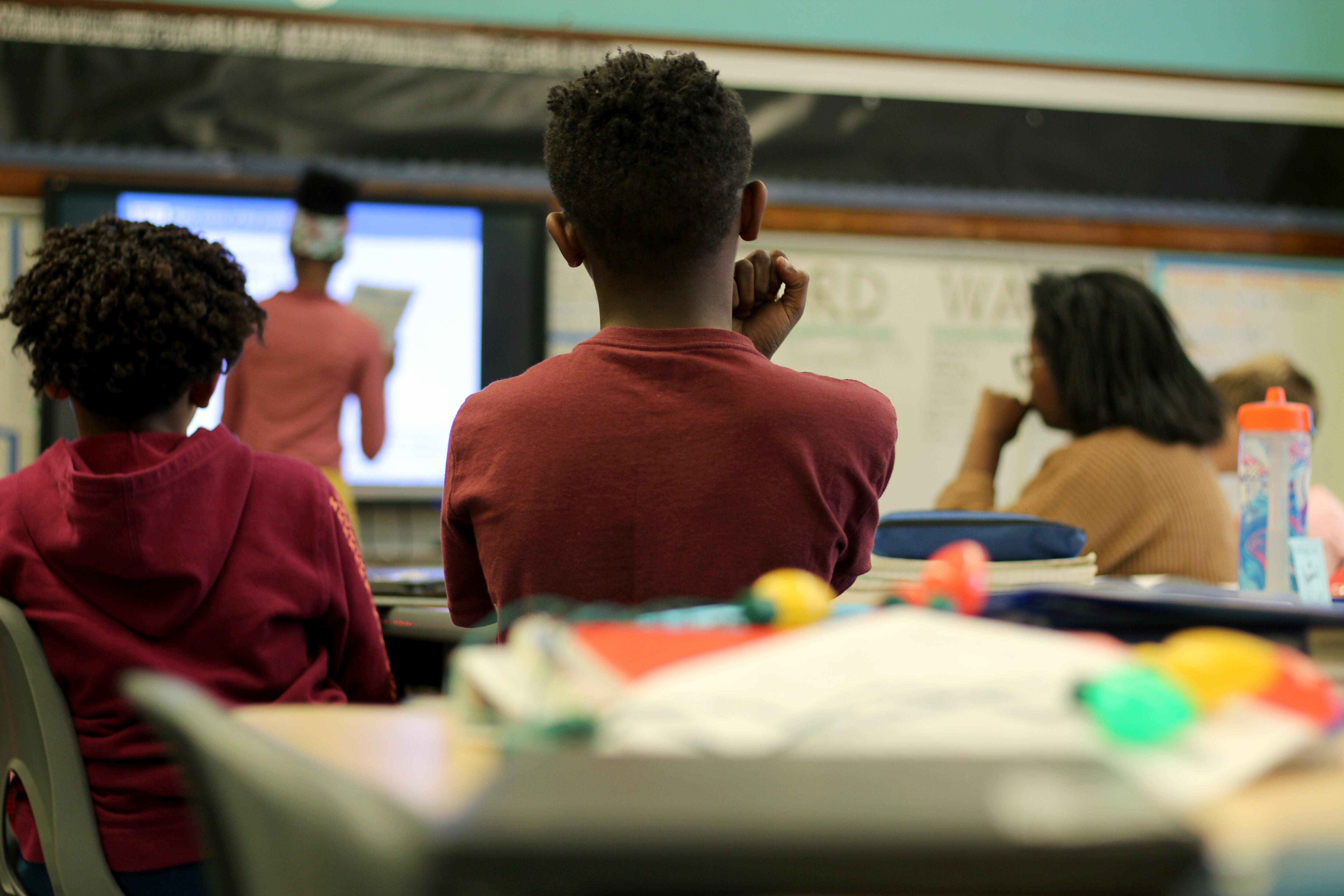 Three students sitting in class face a white board and a projector while their teacher stands at the front of the class.