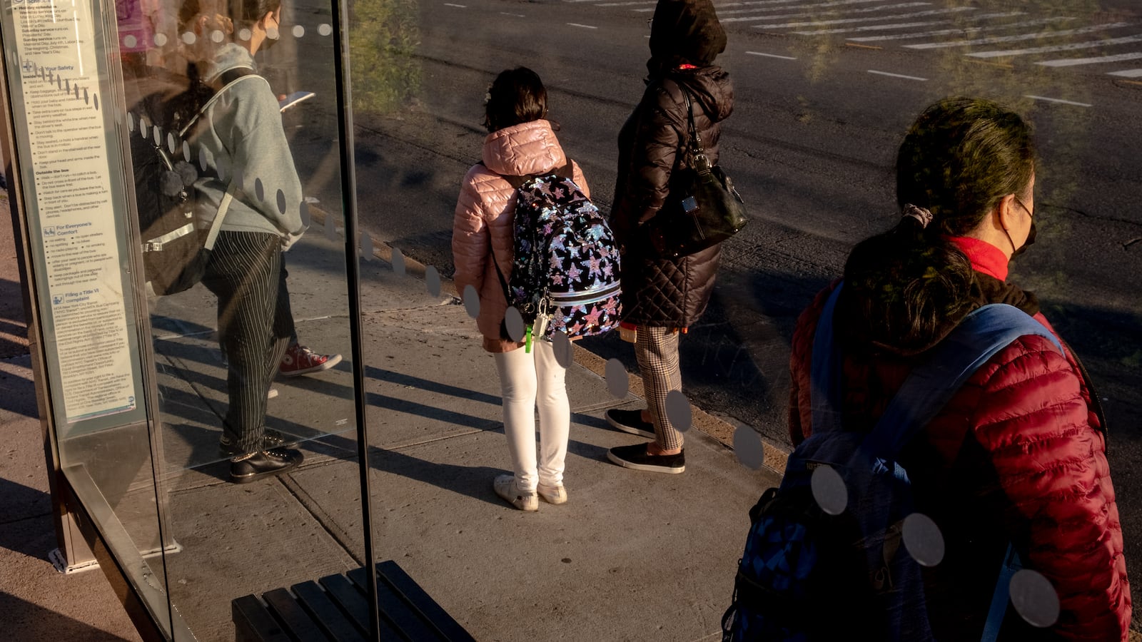 A woman stands with her two daughters and another woman at a city bus stop.