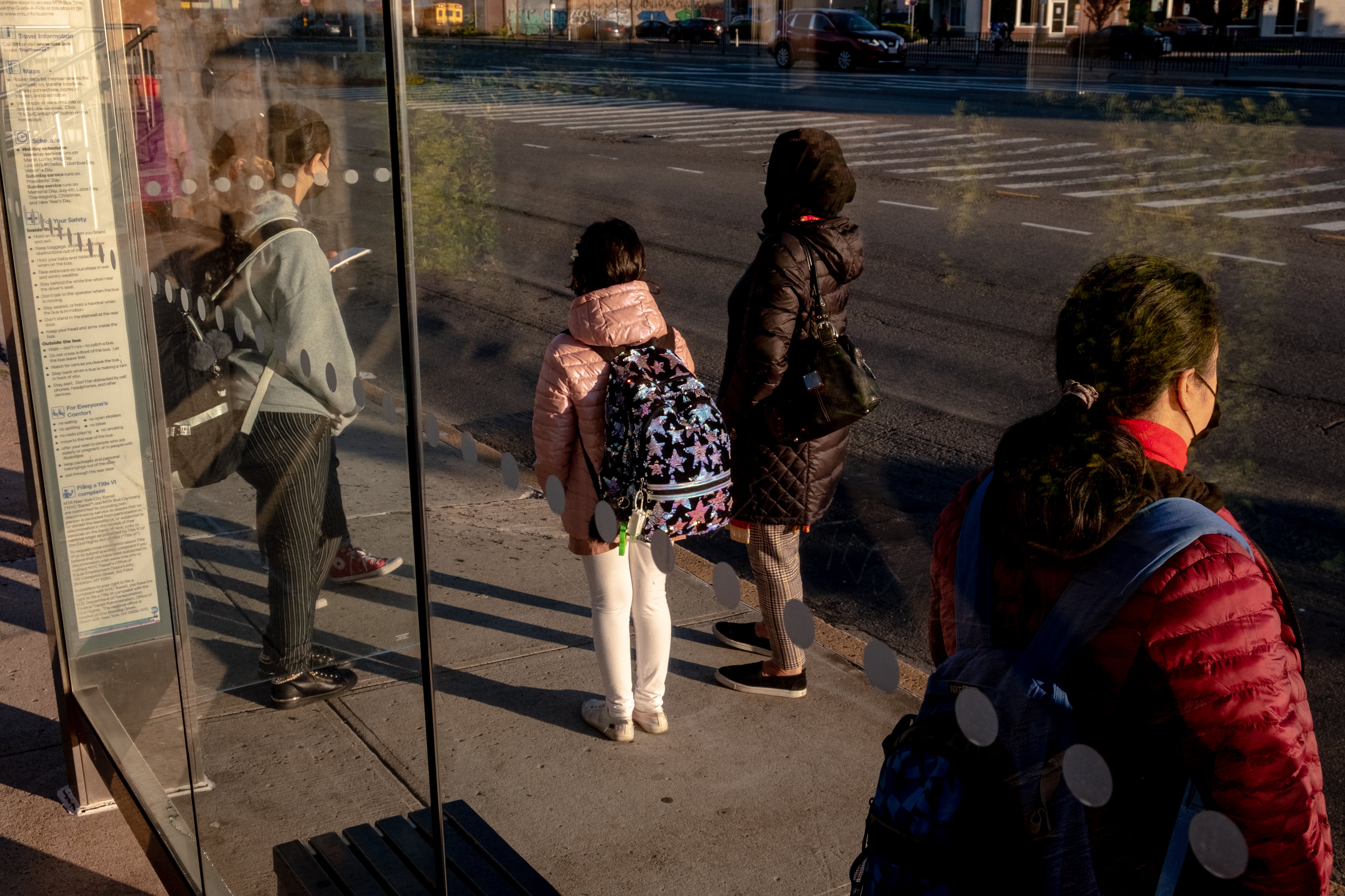 A woman stands with her two daughters and another woman at a city bus stop.