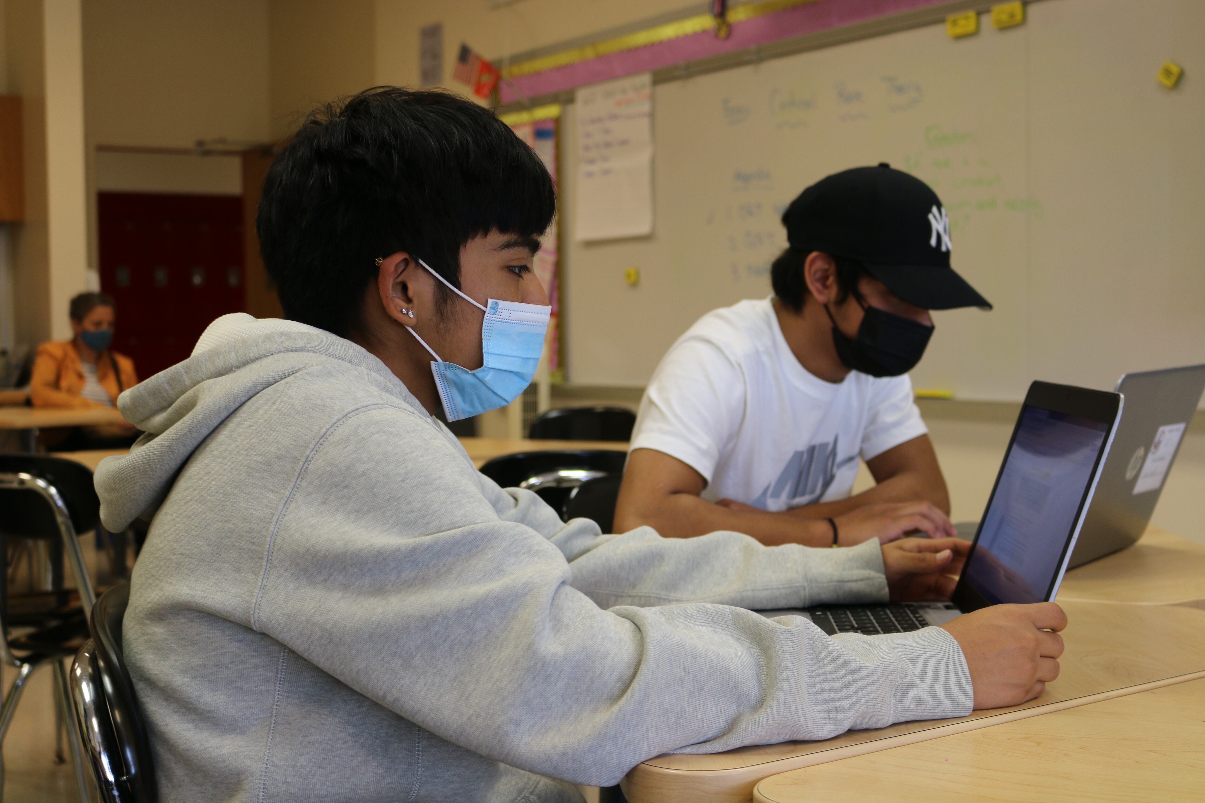 A student wearing a sweatshirt and a mask looks at a laptop on the table in front of him. A second student in a hat can be seen in the background.