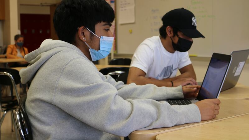 A student wearing a sweatshirt and a mask looks at a laptop on the table in front of him. A second student in a hat can be seen in the background.